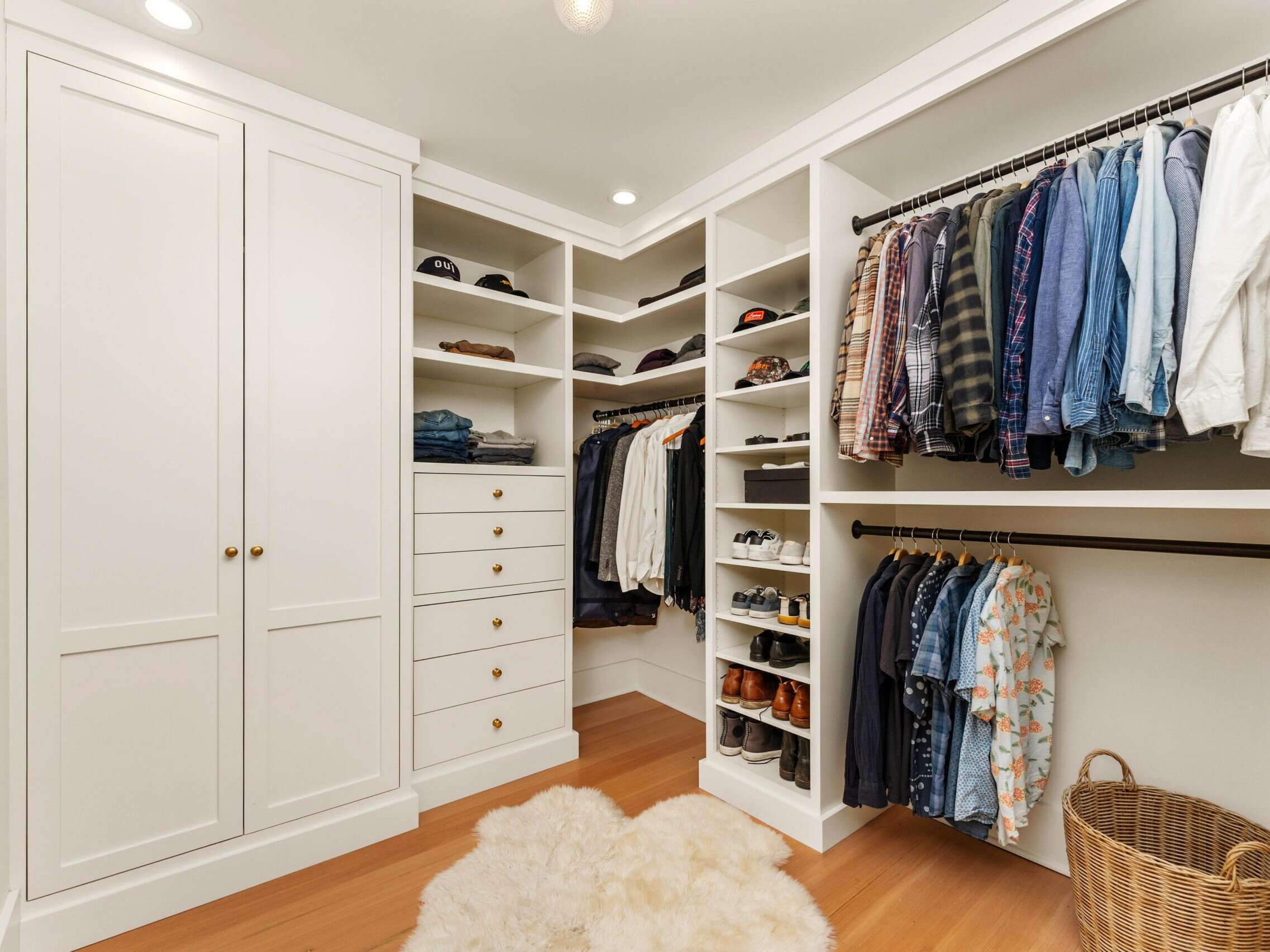 A spacious walk-in closet with white built-in shelves, hanging clothes, drawers, neatly arranged shoes, hats, a wicker laundry basket, and a fluffy white rug on a wooden floor.