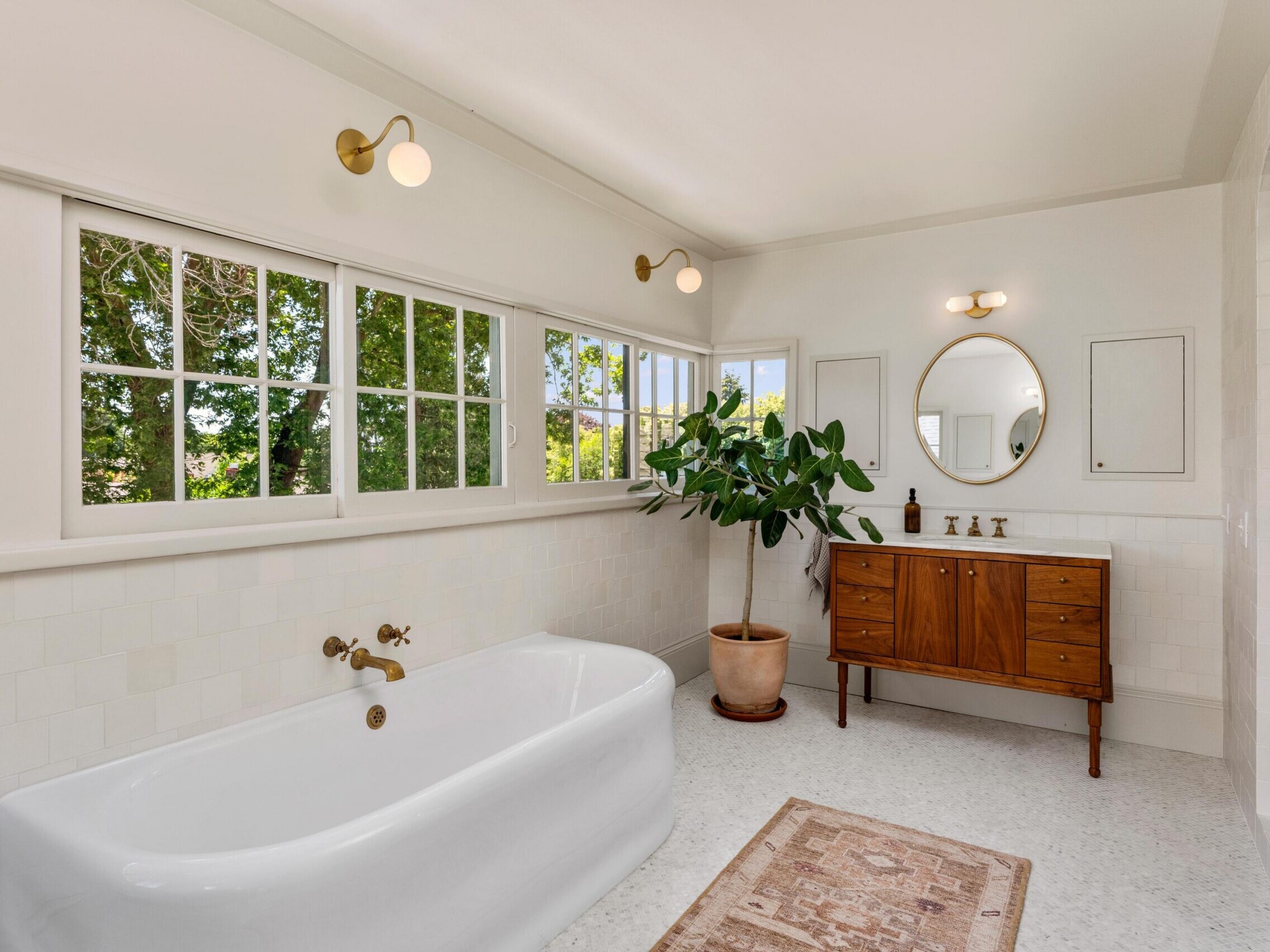 A bright, modern bathroom with a white standalone bathtub, a wooden vanity with a round mirror, gold fixtures, a potted plant, and large windows letting in natural light. A small rug is on the floor.
