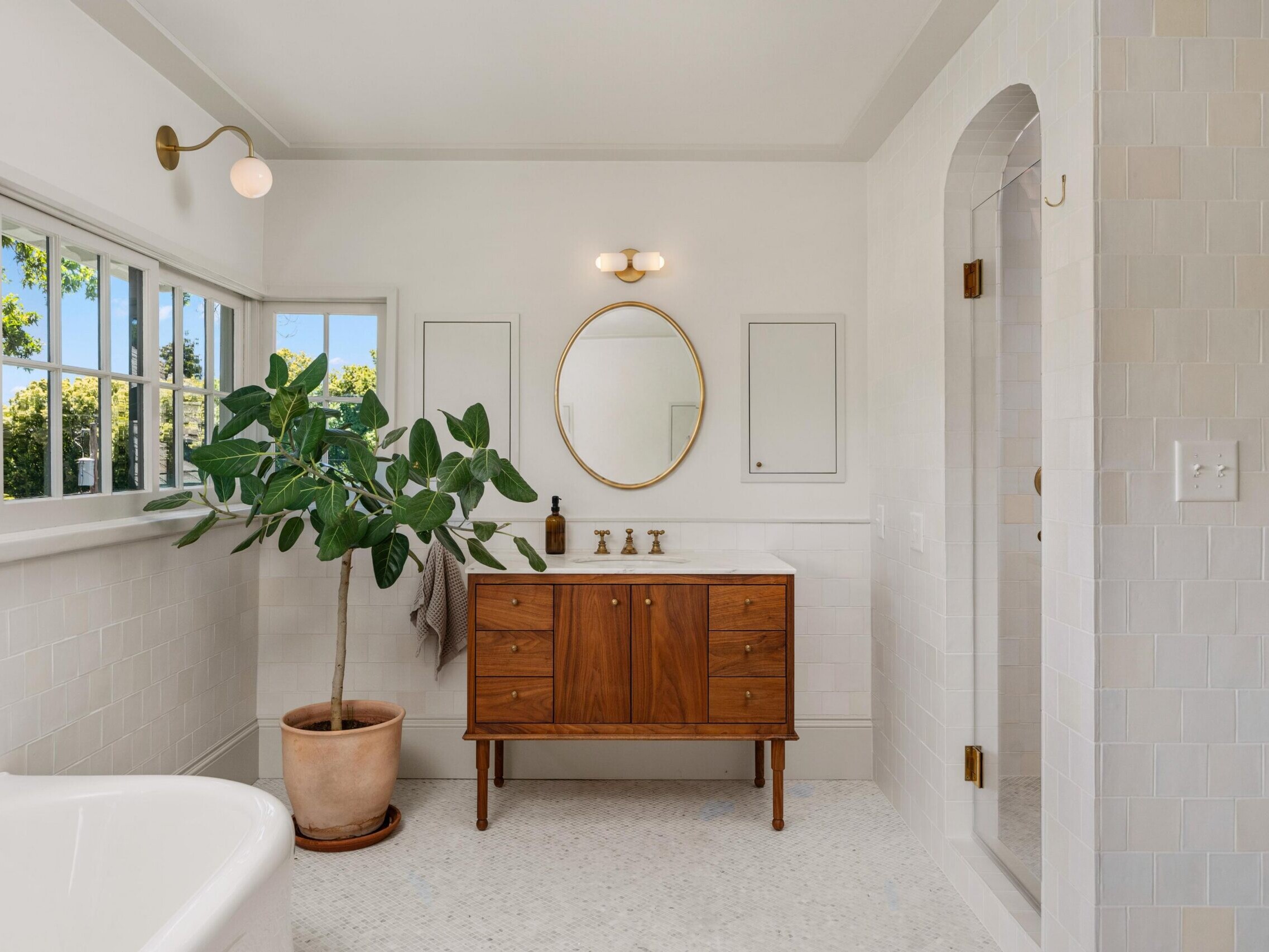 Bright bathroom with white tiled walls, a wooden vanity with a round mirror, gold fixtures, a potted plant, large windows letting in sunlight, and a walk-in shower with a glass door.