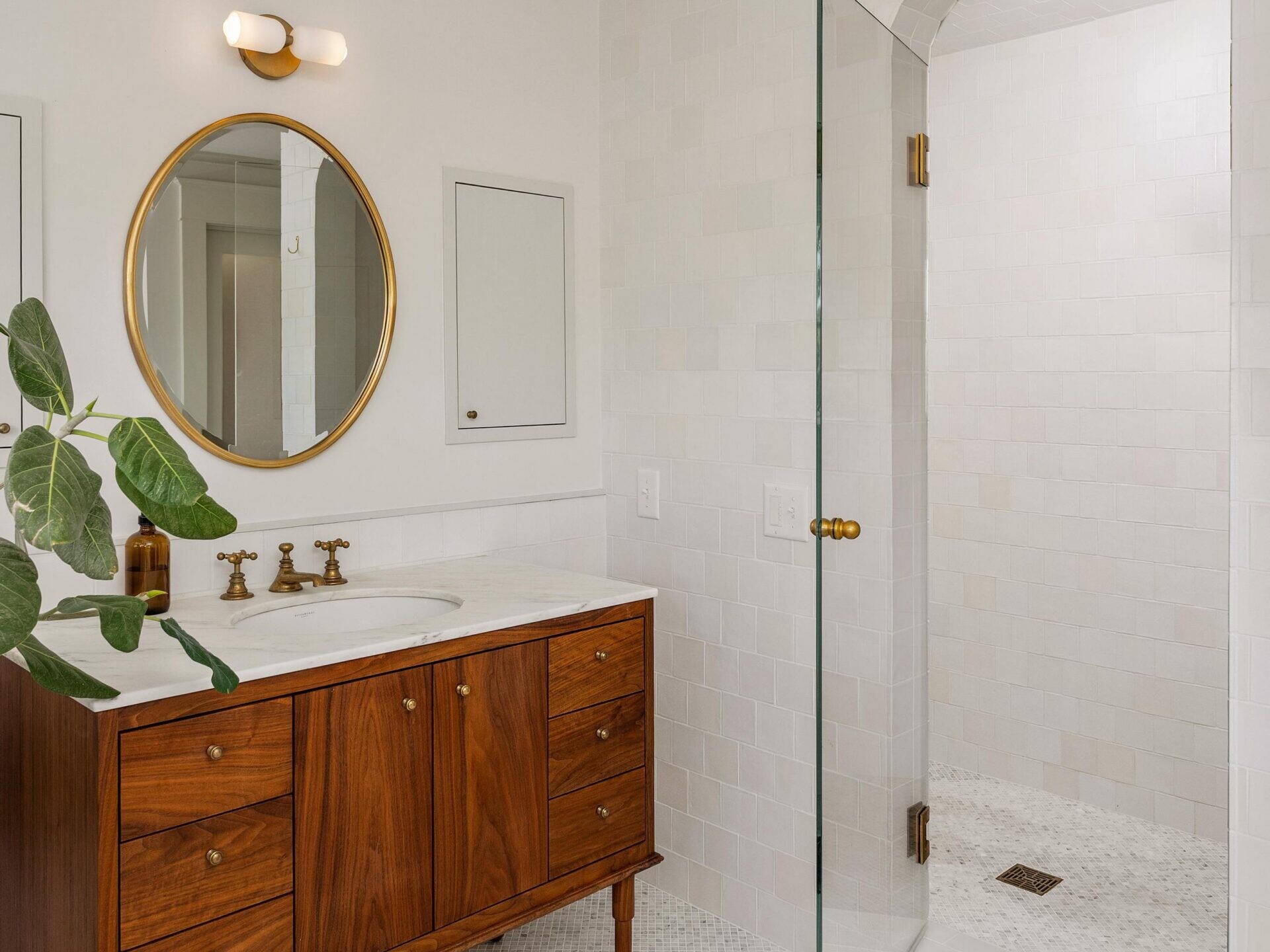 A modern bathroom with a wood vanity, round mirror, white countertop, brass fixtures, a leafy plant, and a glass door leading to a tiled walk-in shower. Walls and floor are light-colored with subtle patterns.