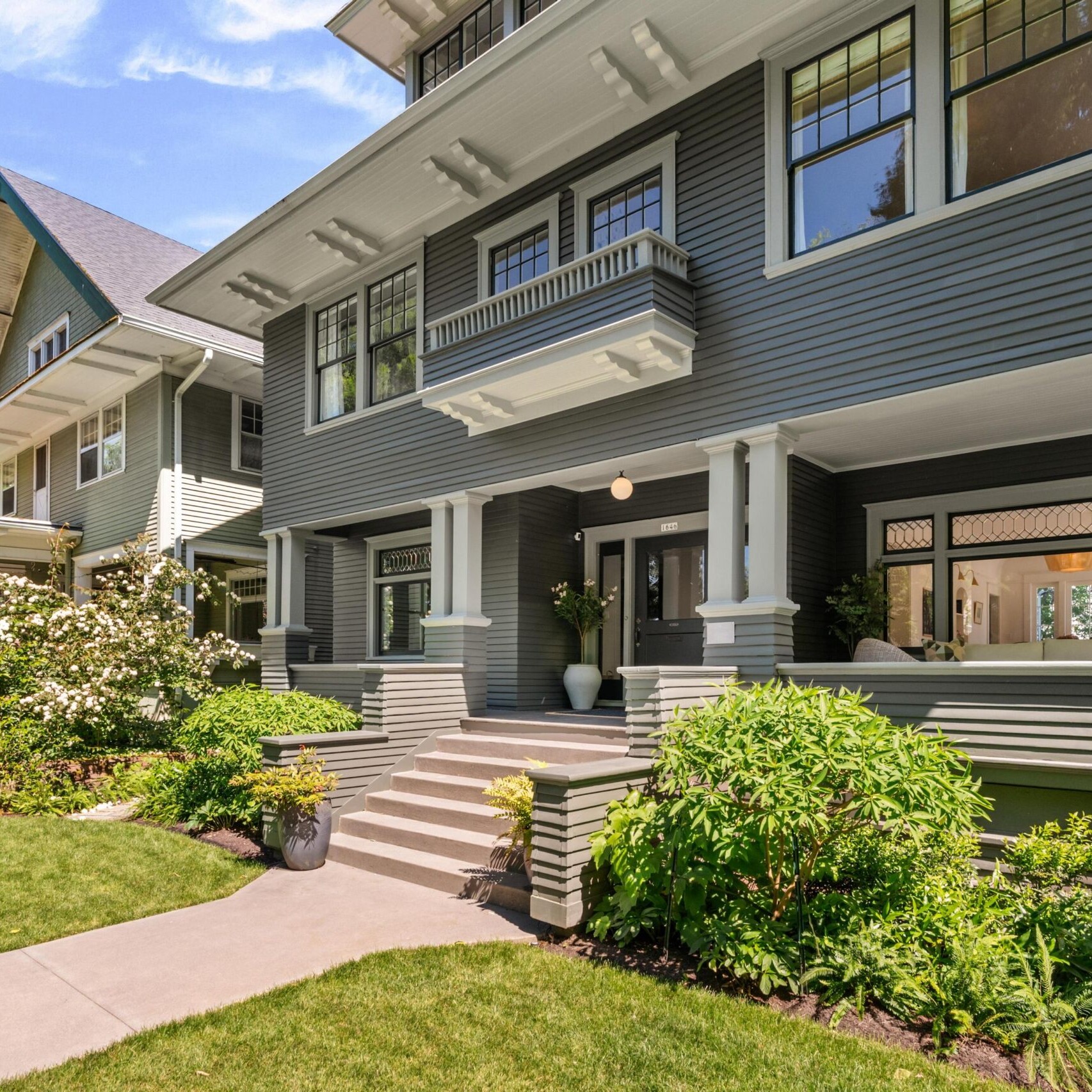 A well-maintained, two-story gray Craftsman-style house with white trim, large windows, and a covered front porch, surrounded by lush green lawn, shrubs, and blooming flowers.