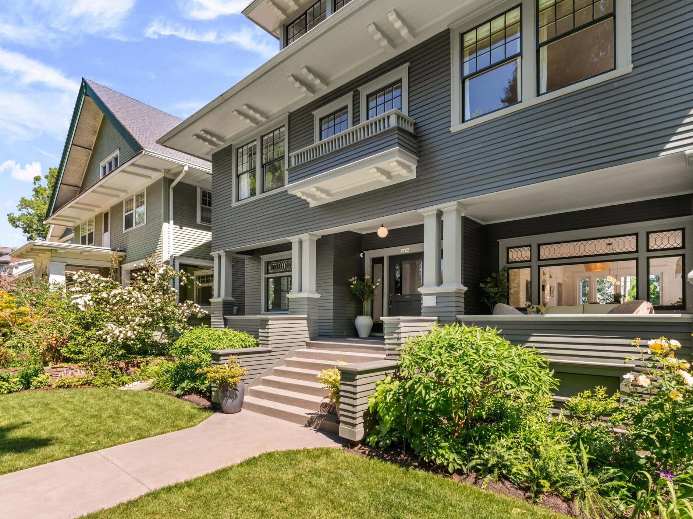 A well-maintained, two-story gray Craftsman-style house with white trim, large windows, and a covered front porch, surrounded by lush green lawn, shrubs, and blooming flowers.