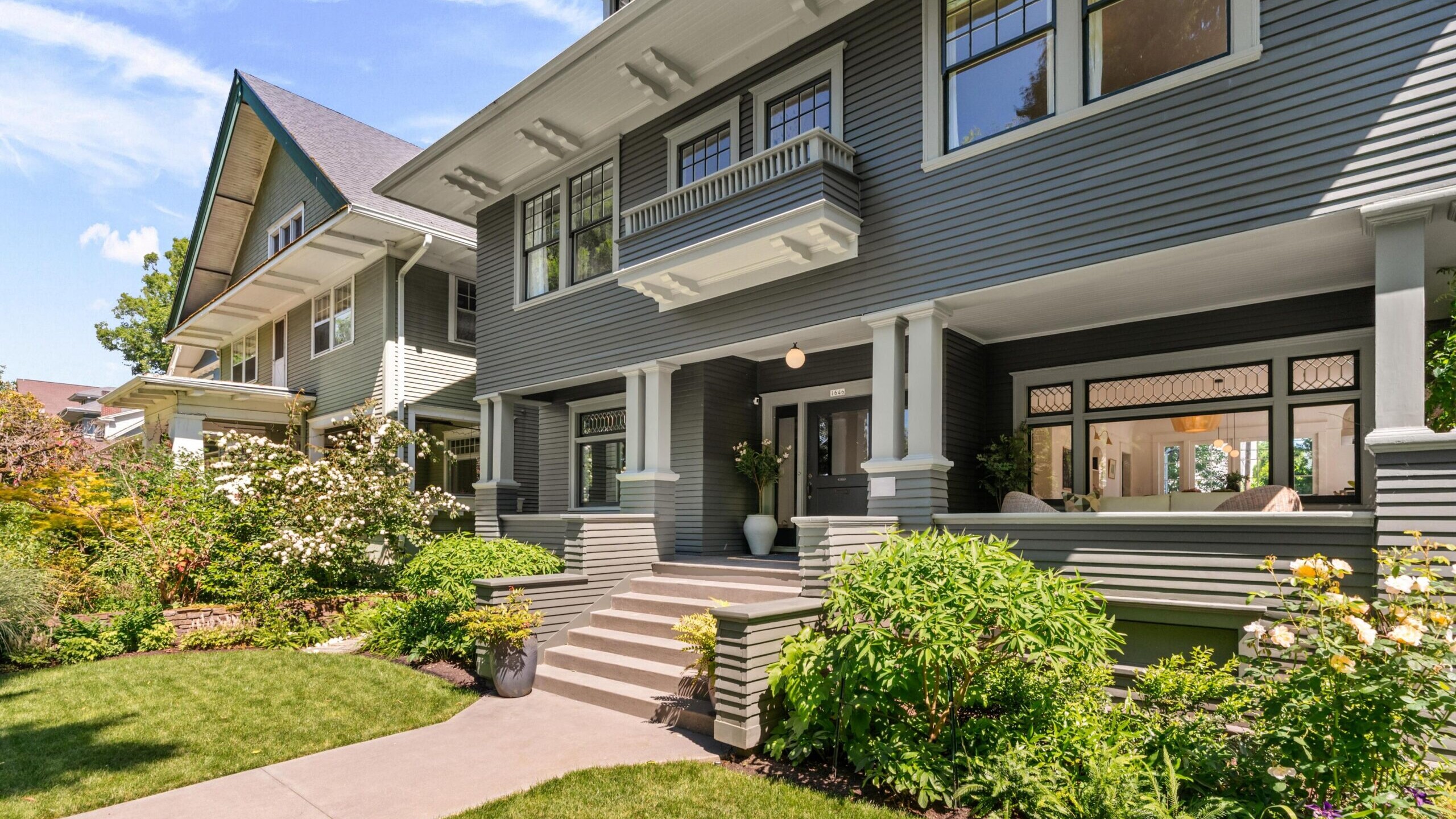 A well-maintained, two-story gray Craftsman-style house with white trim, large windows, and a covered front porch, surrounded by lush green lawn, shrubs, and blooming flowers.