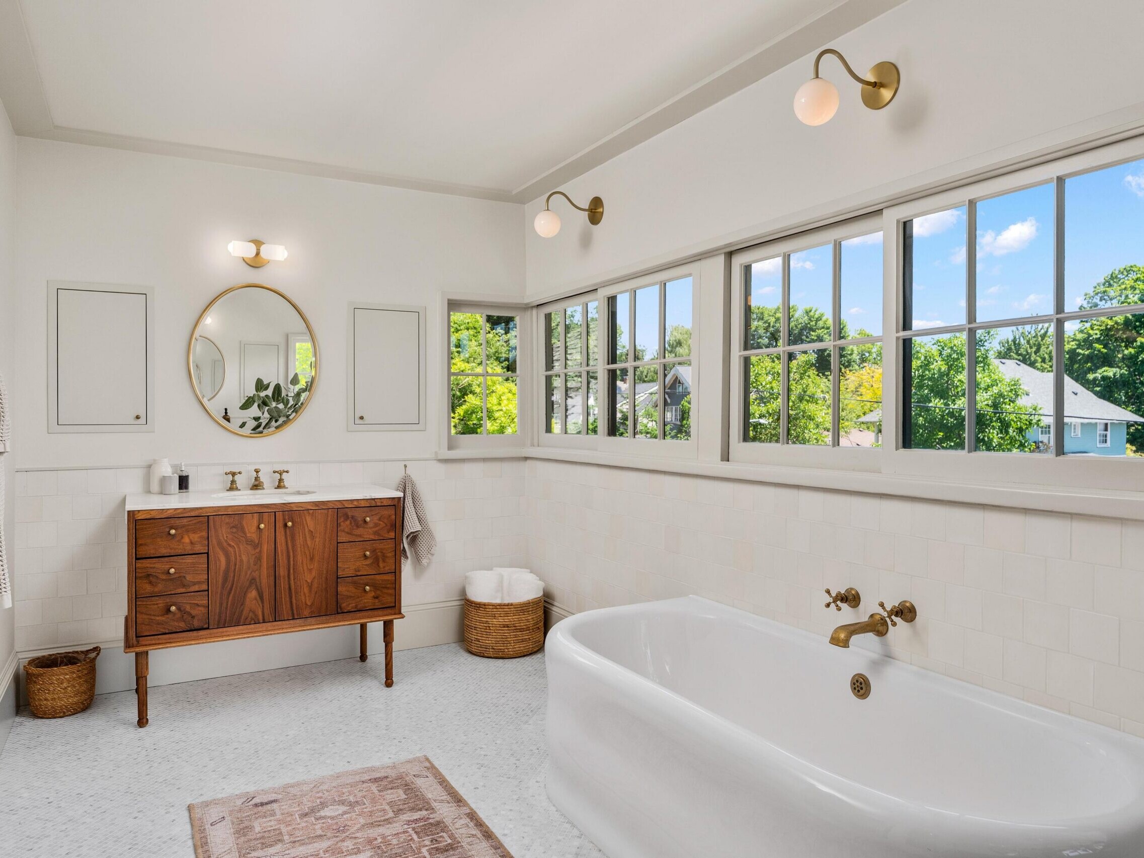 A bright, spacious bathroom with a large white bathtub, wooden vanity with a round mirror, brass fixtures, wall sconces, a woven basket, and wide windows letting in natural light with a view of greenery outside.