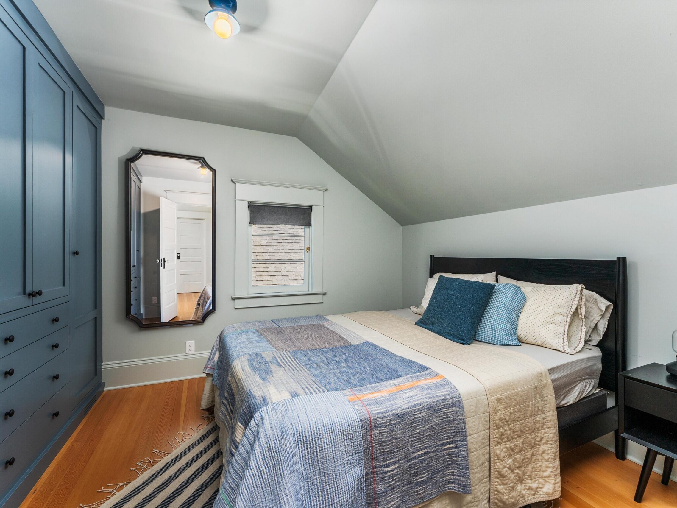 A cozy bedroom with a black bed, blue and beige bedding, blue built-in wardrobes, a black nightstand with a lamp, a striped rug, and a large wall mirror reflecting a hallway.