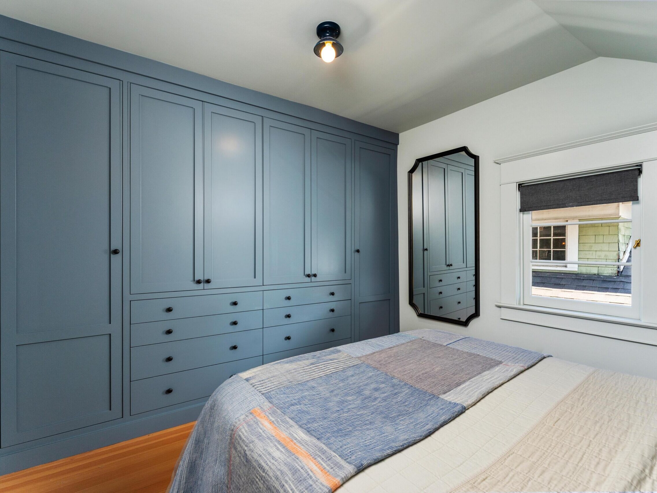 A bedroom with a large blue built-in wardrobe, a bed with a patchwork quilt, a black-framed mirror on the wall, and a window with a dark gray Roman shade. The room has white walls and wooden flooring.