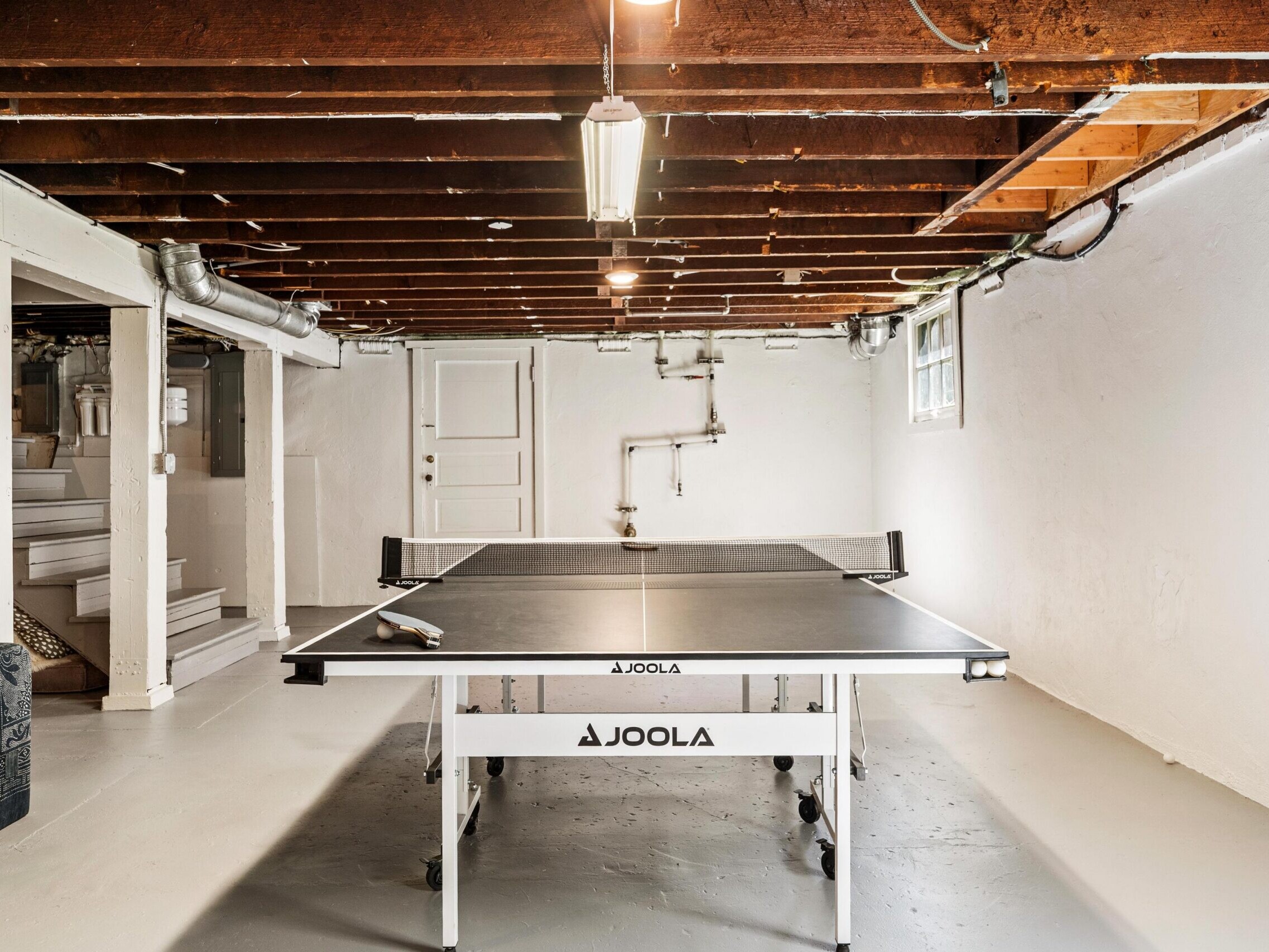 A basement with exposed wooden beams and white walls features a JOOLA ping pong table in the center, with paddles on the table, and a small window and steps leading upstairs in the background.