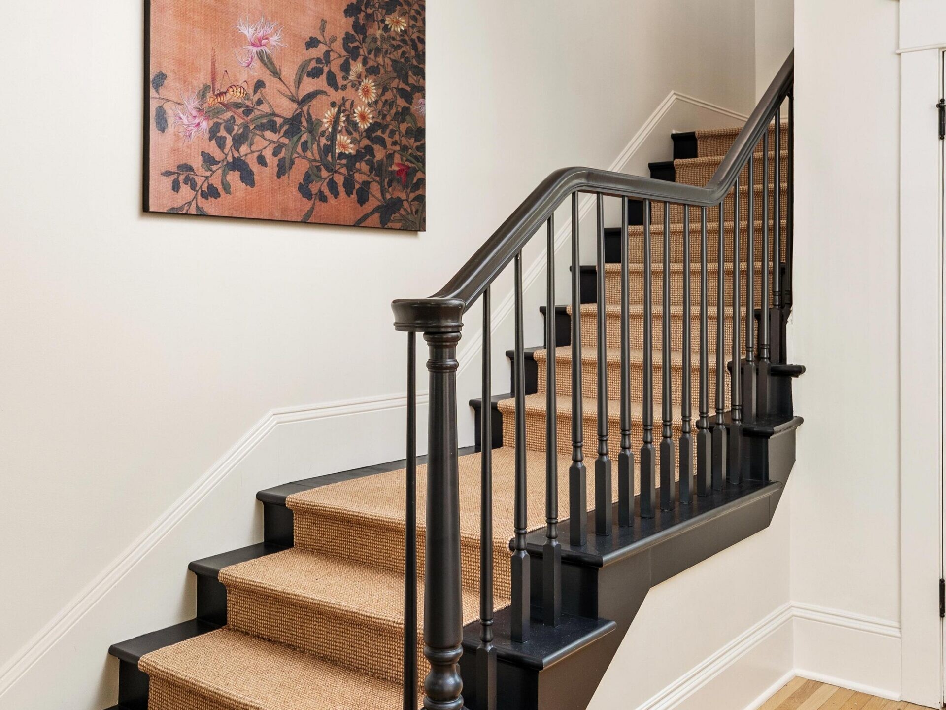 A staircase with tan carpet and black railings leads up in a bright hallway with light wood floors. A large floral painting with a brown background hangs on the wall at the turn of the stairs.