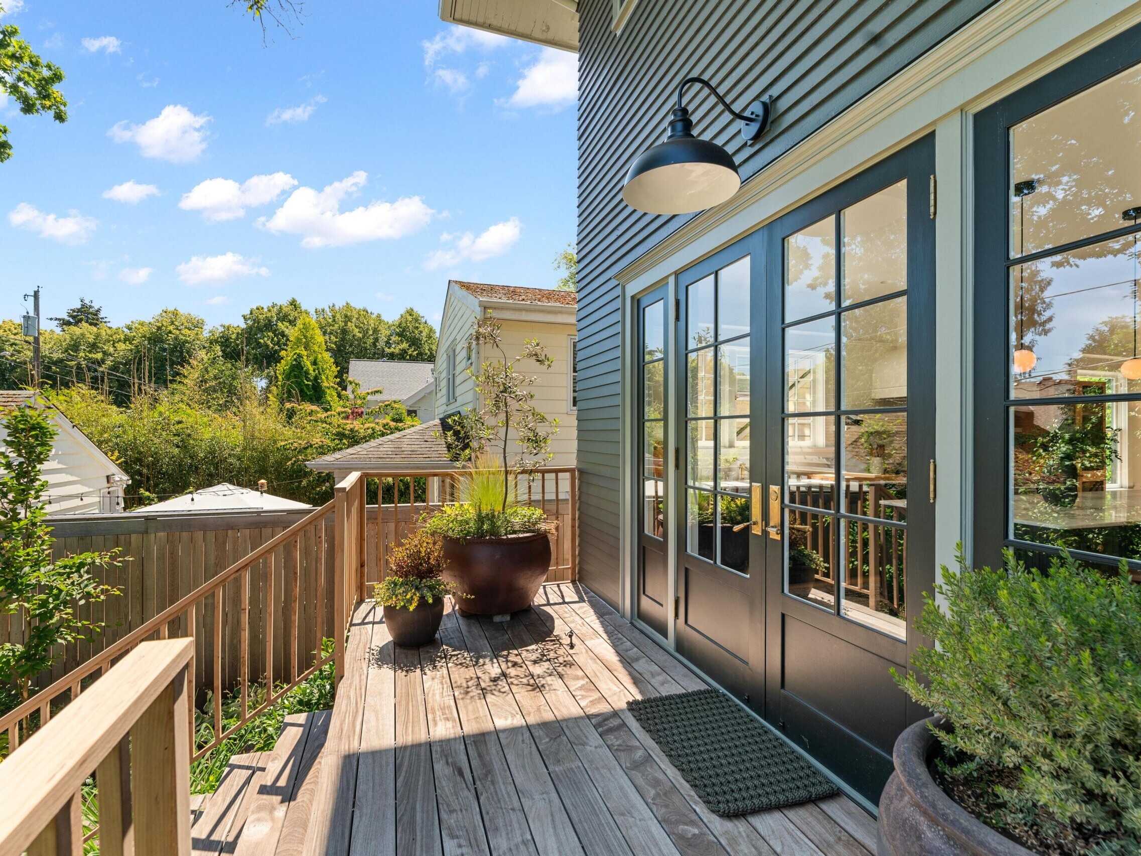 A wooden deck with potted plants and a mat sits outside dark double doors, attached to a house with gray siding. A fence, trees, and neighboring houses are visible under a bright, blue sky with clouds.