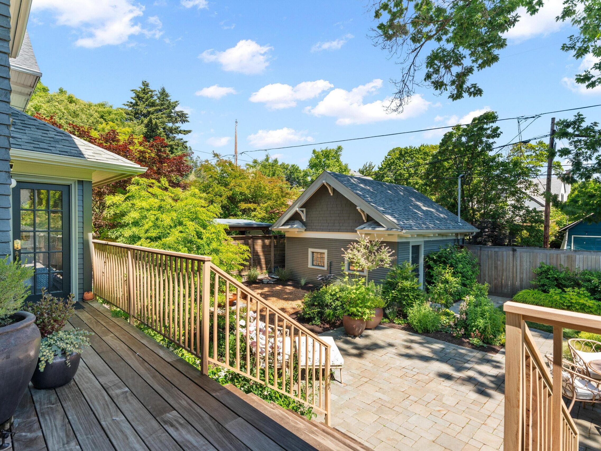 View from a wooden deck with railing overlooking a landscaped backyard, potted plants, a stone patio, outdoor seating, and a small gray outbuilding, surrounded by trees and a wooden fence under a blue sky.