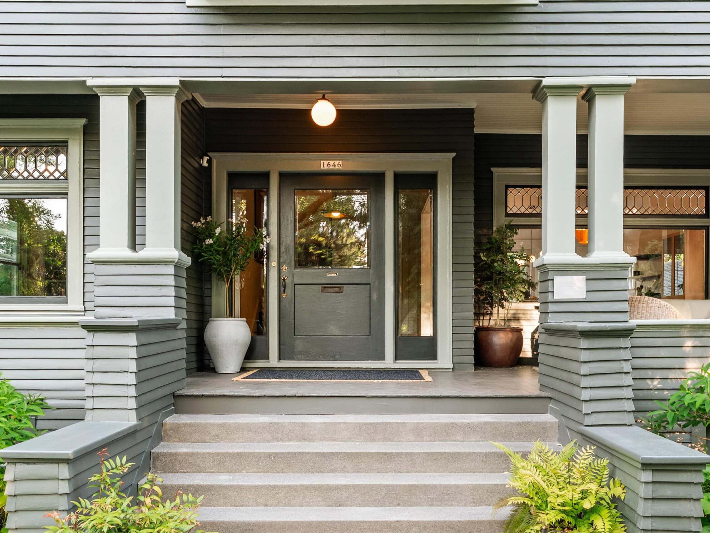 Front porch of a gray house with wide steps, two potted plants flanking the door, a doormat, and a round light above the entrance. Large windows and greenery surround the entryway.