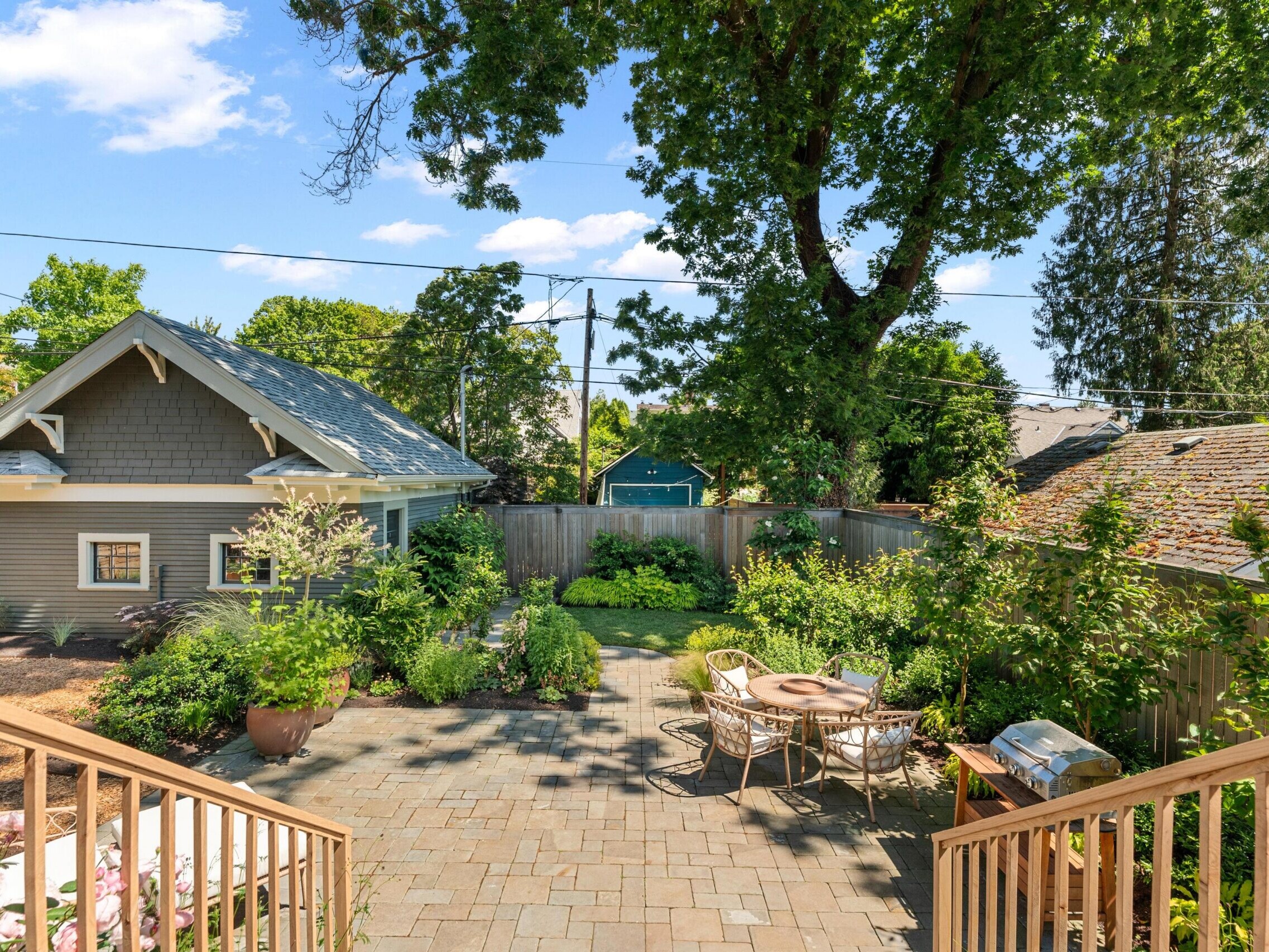 A sunny backyard with a paved patio, outdoor dining table, barbecue grill, lush green plants, a large tree, and a small house or shed surrounded by a wooden fence.