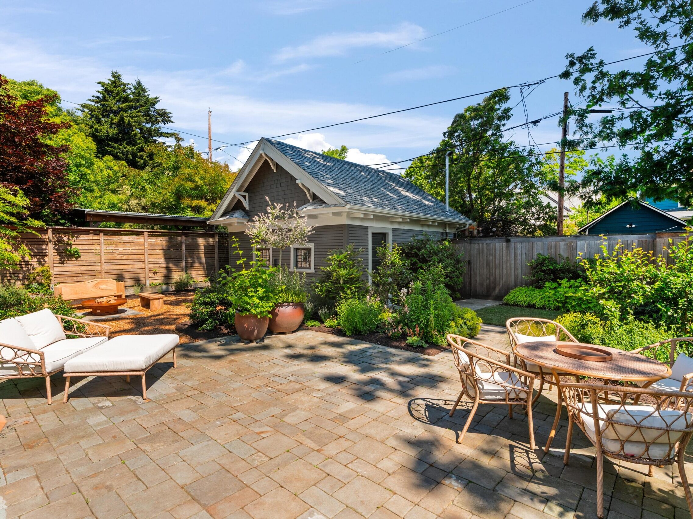 Sunny backyard with stone patio, white cushioned lounge chairs, a round wicker dining table with chairs, potted plants, lush green trees, and a small shed with a gray roof in the background.