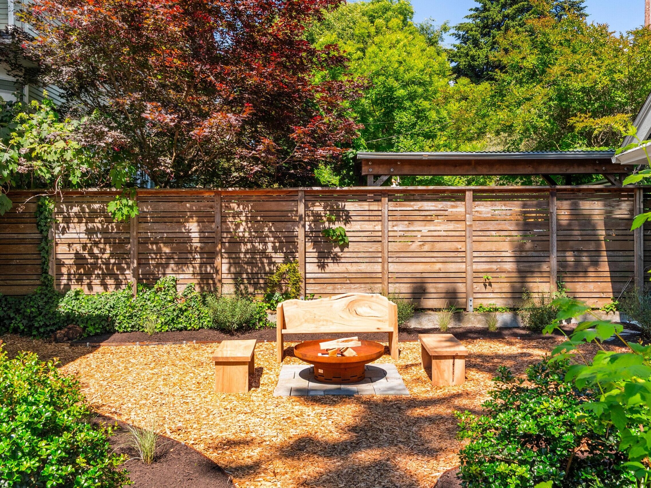 Sunny backyard with a wooden fence, a fire pit, and wooden benches on a mulch-covered area, surrounded by trees and green plants.