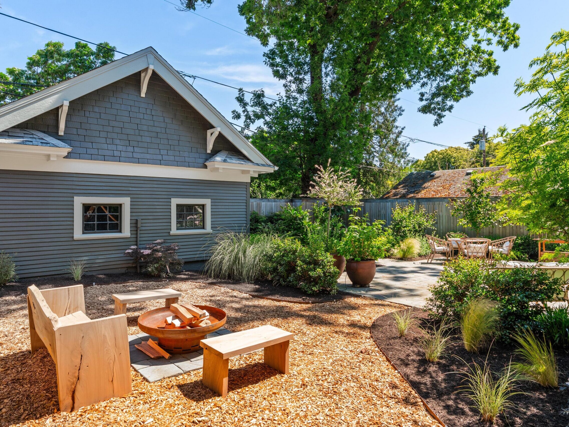 A backyard with a wooden shed, fire pit surrounded by wooden benches, lush greenery, mulch ground cover, patio area with chairs, and trees in the background on a sunny day.