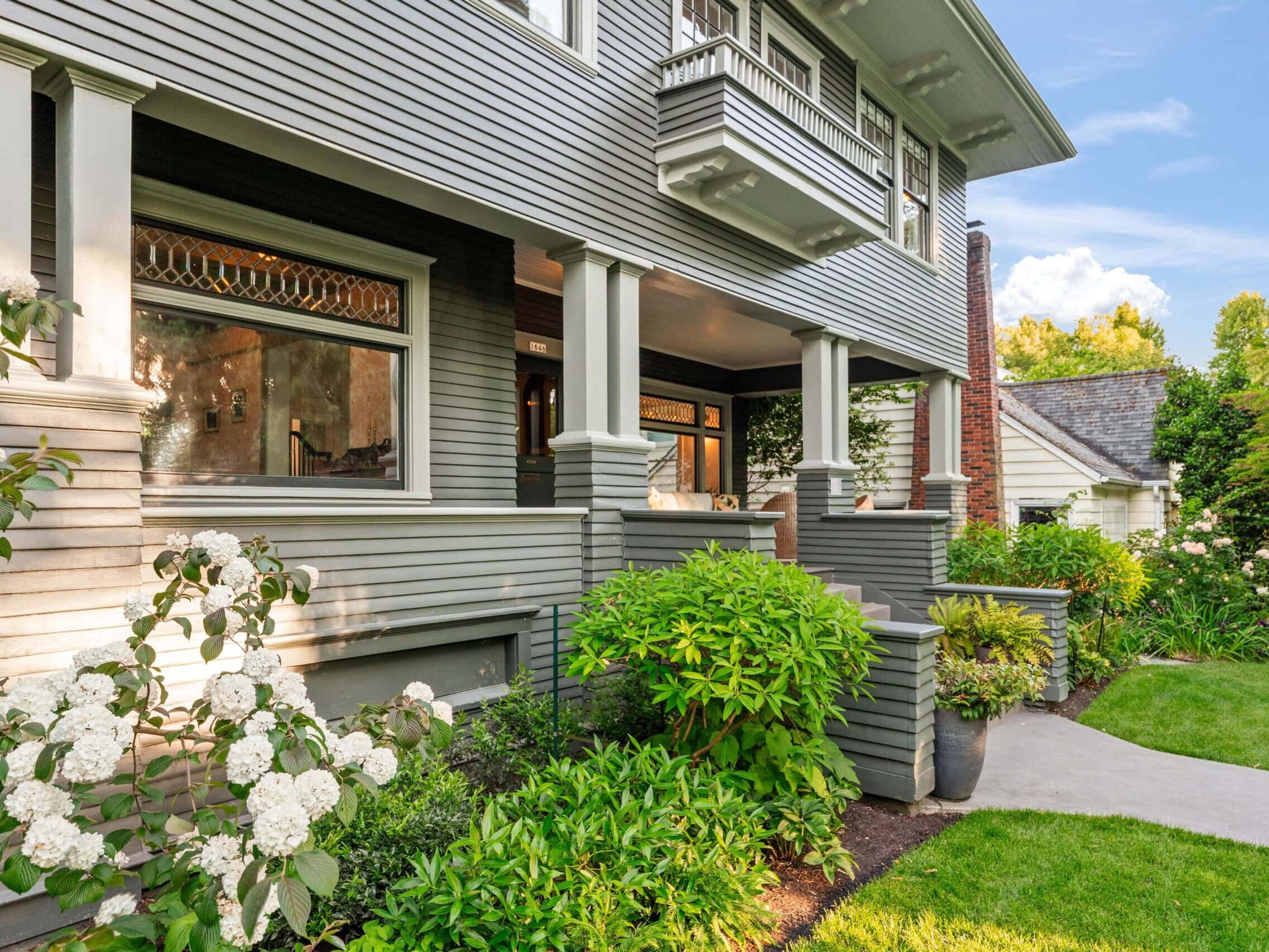 A gray two-story house with large windows, white trim, and a covered front porch, surrounded by lush green shrubs, flowering plants, and a manicured lawn on a sunny day.