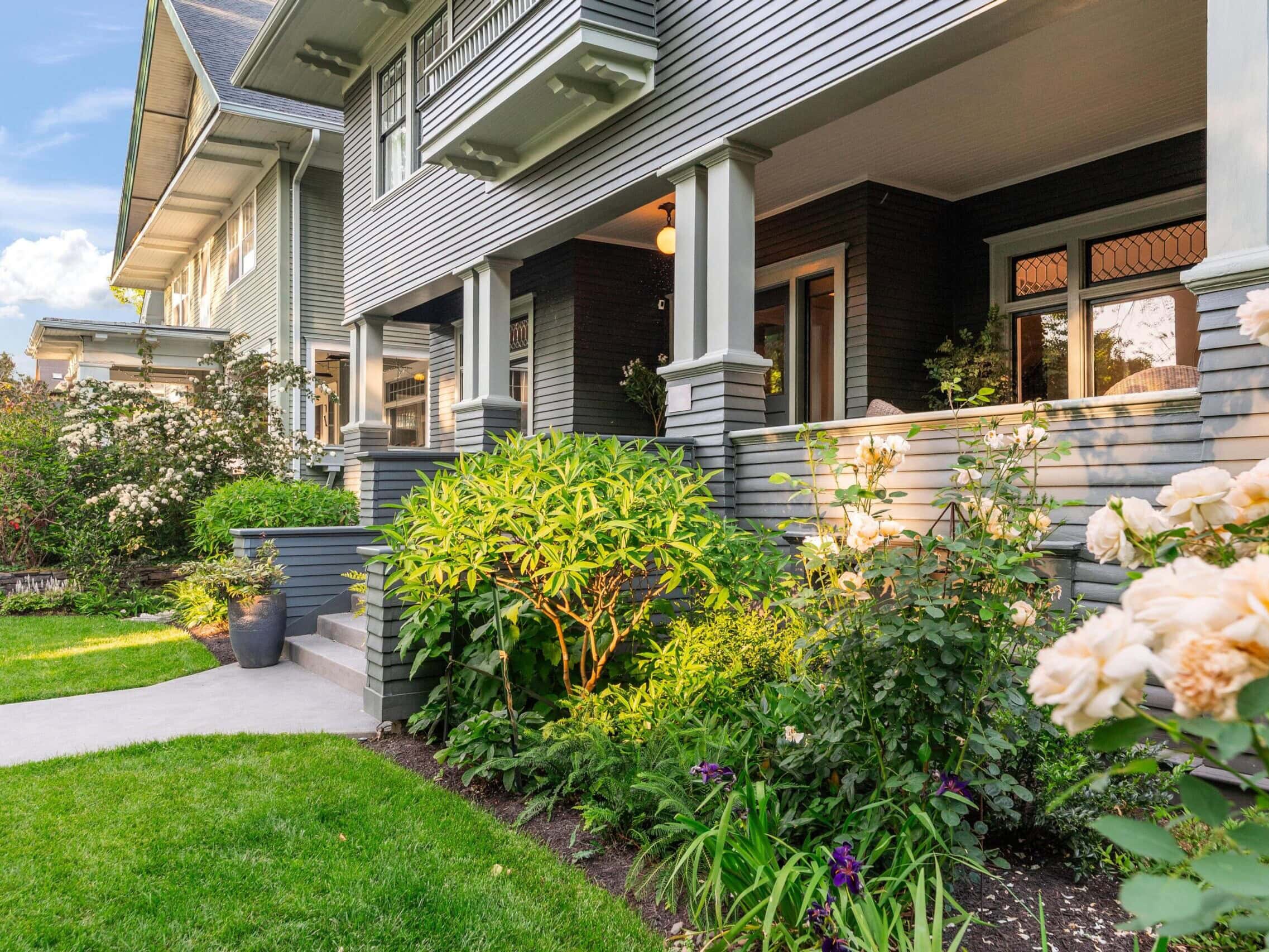 A well-maintained house with gray siding, a large porch, and white trim, surrounded by a lush green lawn, flowering shrubs, and blooming rose bushes on a sunny day.