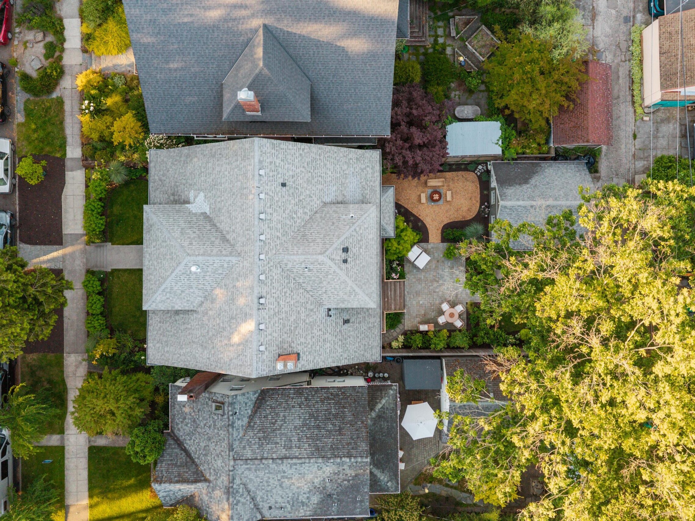 Aerial view of a suburban neighborhood showing several houses with gray roofs, green trees, gardens, sidewalks, parked cars, and a backyard patio with outdoor furniture.