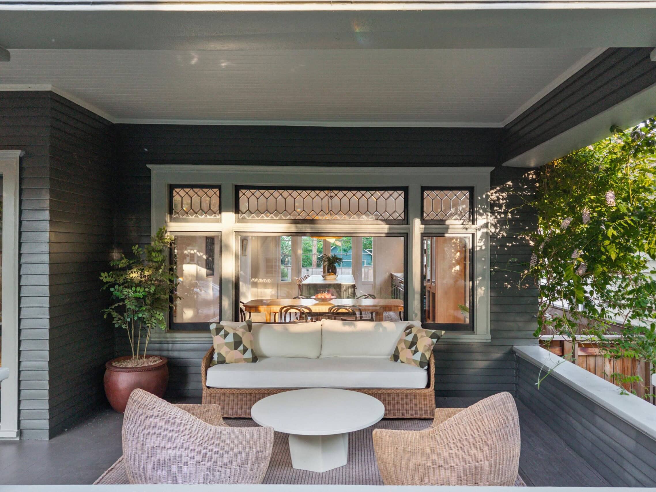 A cozy porch with a cushioned sofa, two woven chairs, and a round white table. Potted plants decorate the space, and large windows show a dining area inside the house.