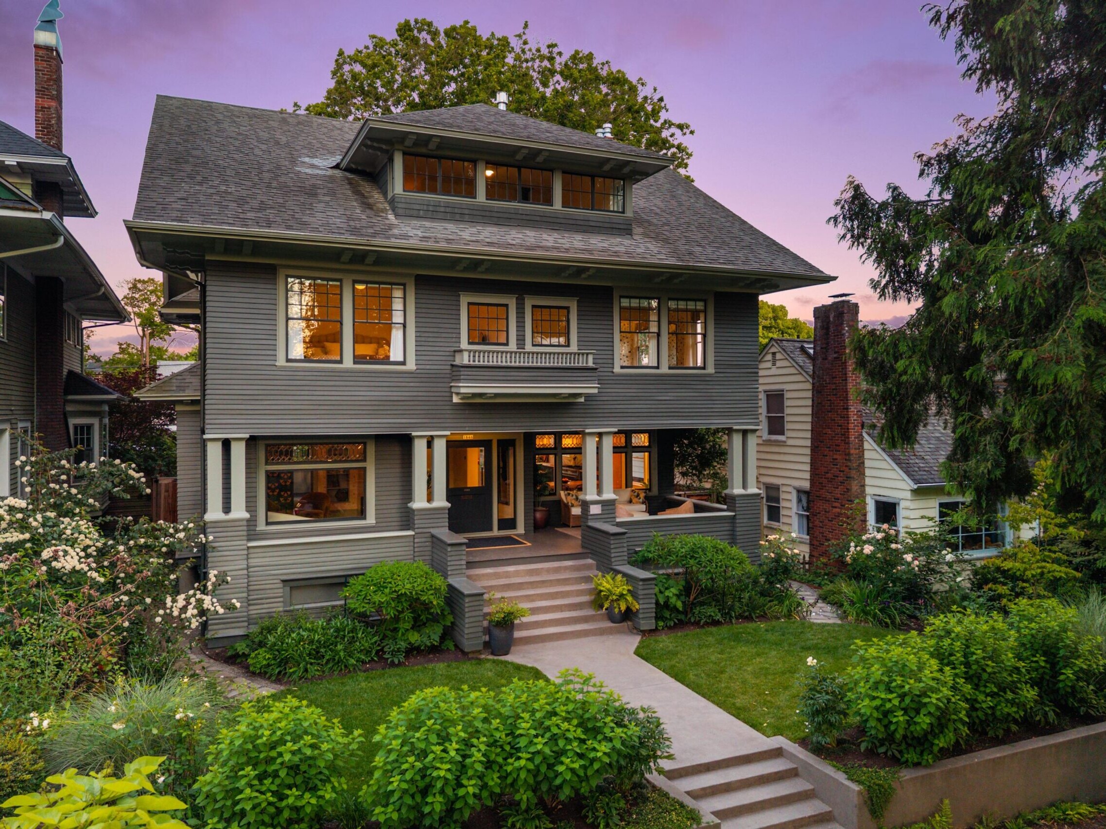 A two-story gray Craftsman-style house with large windows, a covered front porch, and manicured landscaping, photographed at sunset with a pink and purple sky in the background.