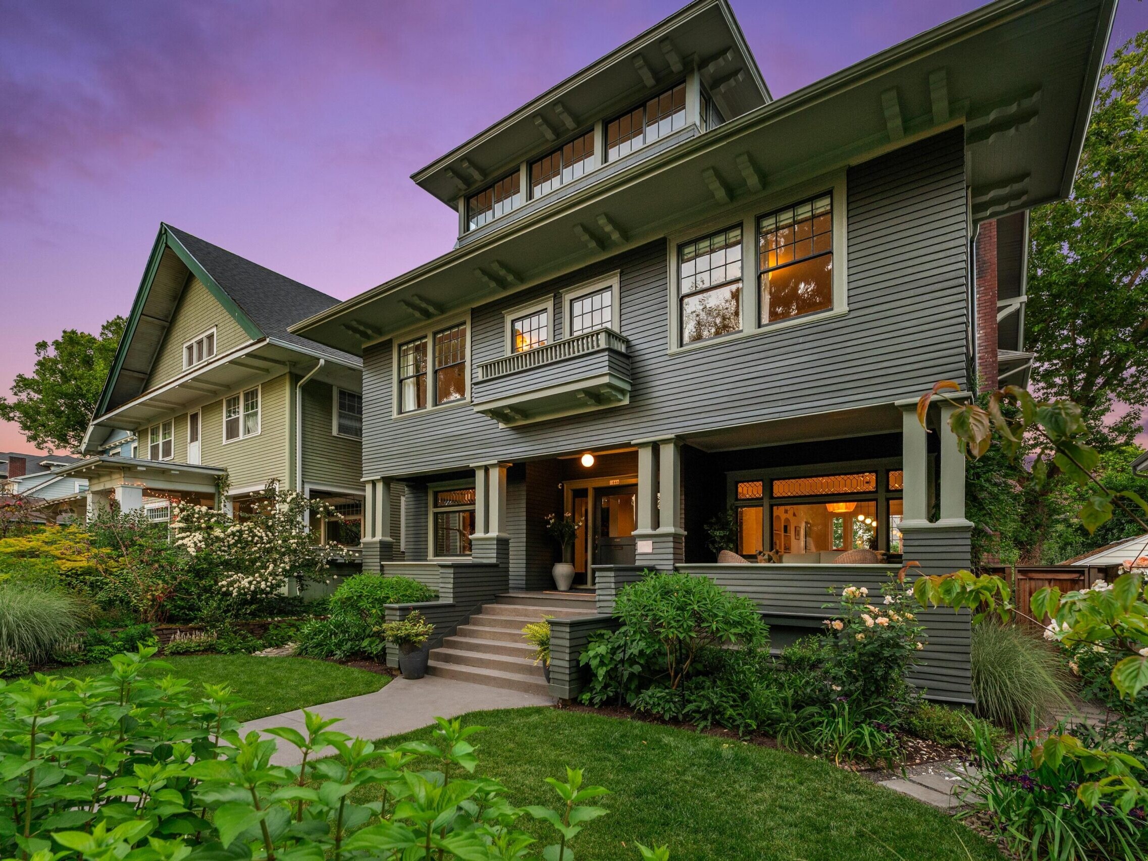 A large, two-story green Craftsman-style house with a wide front porch and landscaped yard, pictured at sunset with a purple sky. Neighboring houses and lush greenery surround the home.