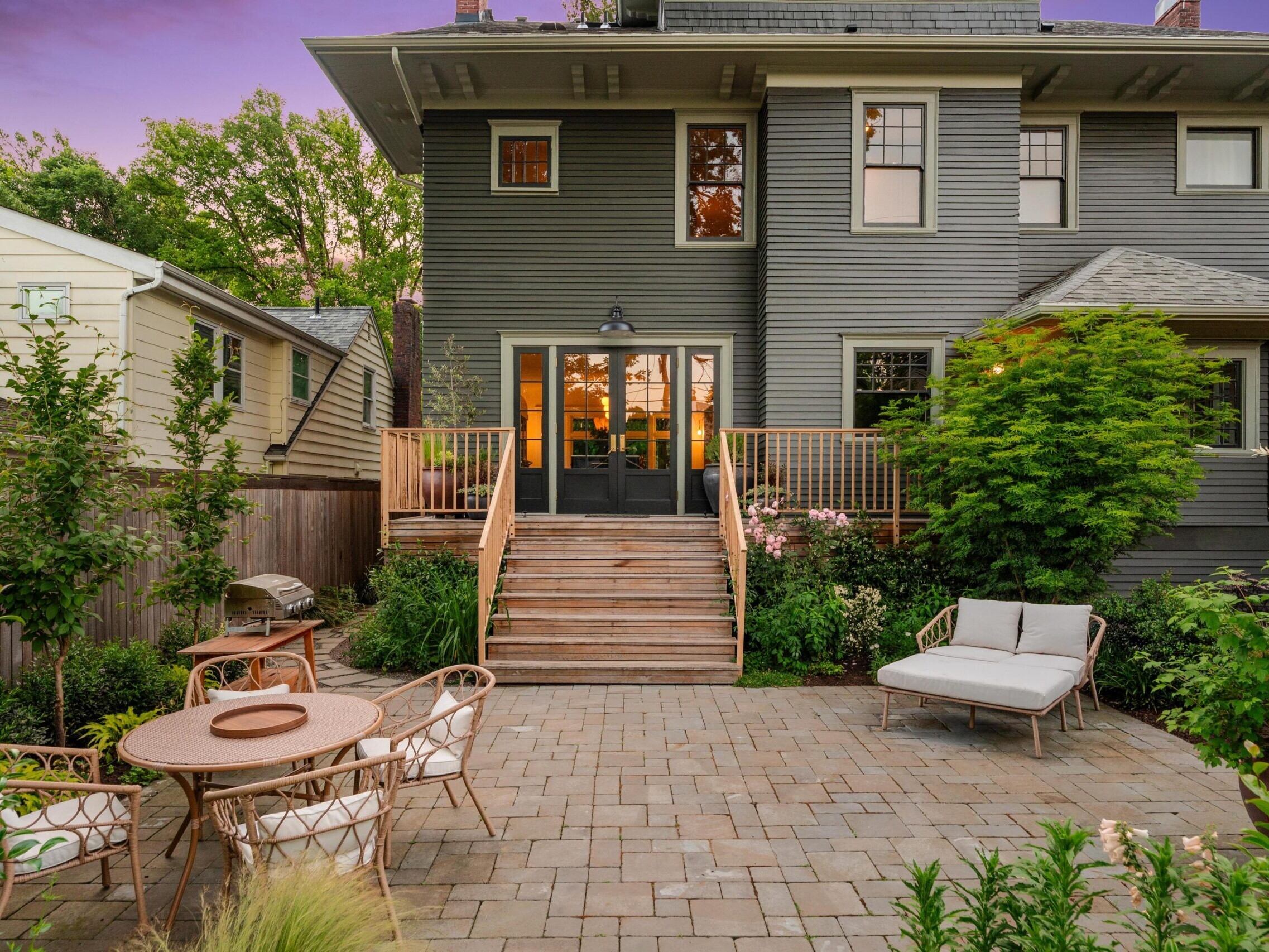 Backyard patio with stone pavers, outdoor dining set, cushioned lounge chair, greenery, and wooden stairs leading to a gray two-story house in the evening with warm light inside.
