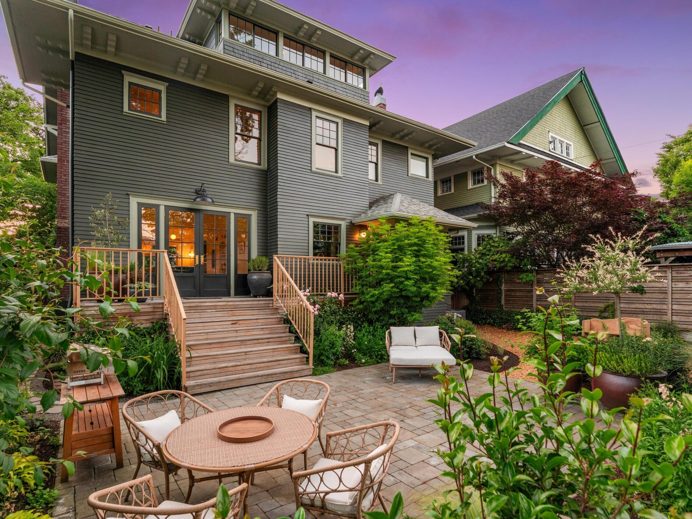 A spacious backyard with a brick patio, outdoor dining set, lounge chair, lush greenery, and a large gray house with wide stairs leading to double doors at sunset.