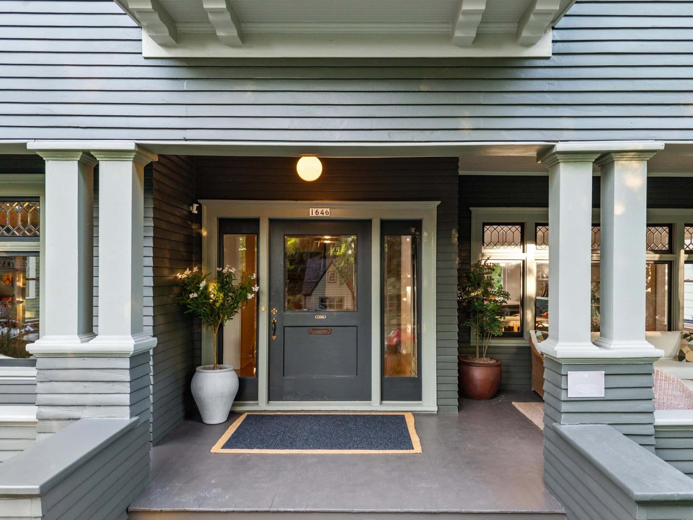 Front porch of a house with gray siding, a blue door with glass panels, two large potted plants, and windows on either side. A doormat and overhanging light are present.