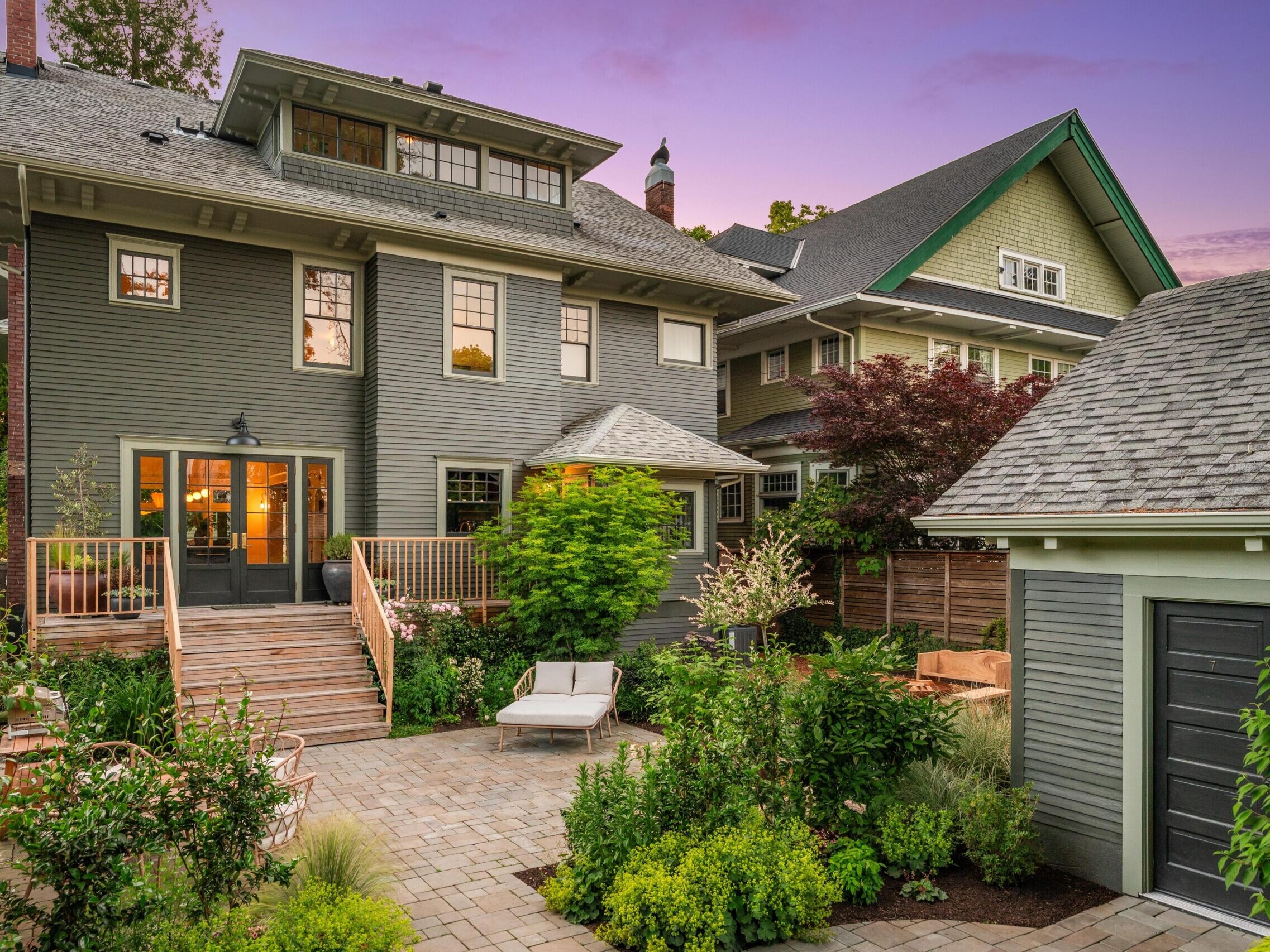 A charming two-story gray house with a back porch, surrounded by lush greenery and a landscaped patio, at sunset. Another house and a garage are visible nearby, with a purple and pink sky in the background.