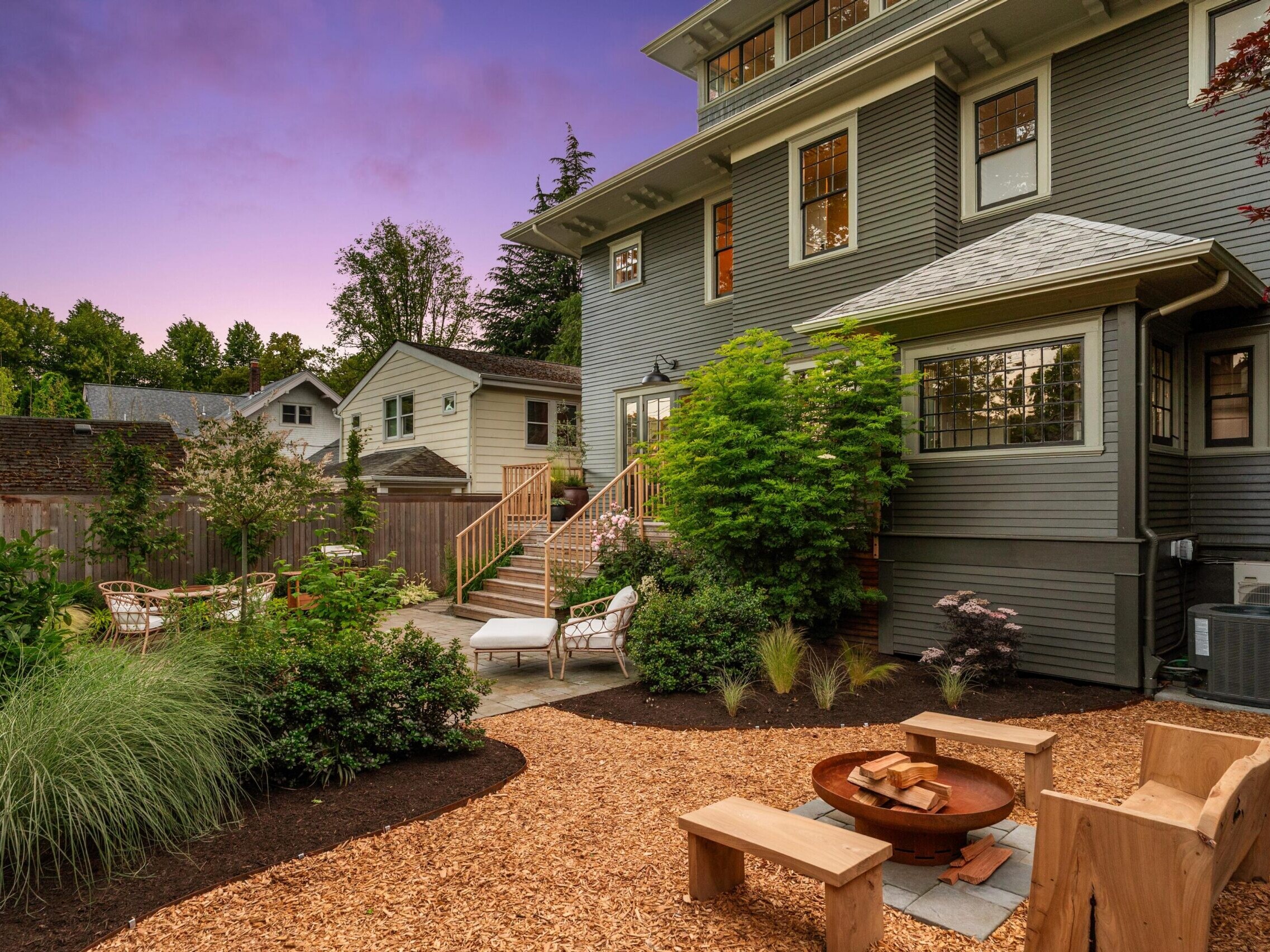 A landscaped backyard at dusk with a wooden bench and fire pit, a gravel seating area, lush green plants, and a gray house with multiple windows and a patio in the background.