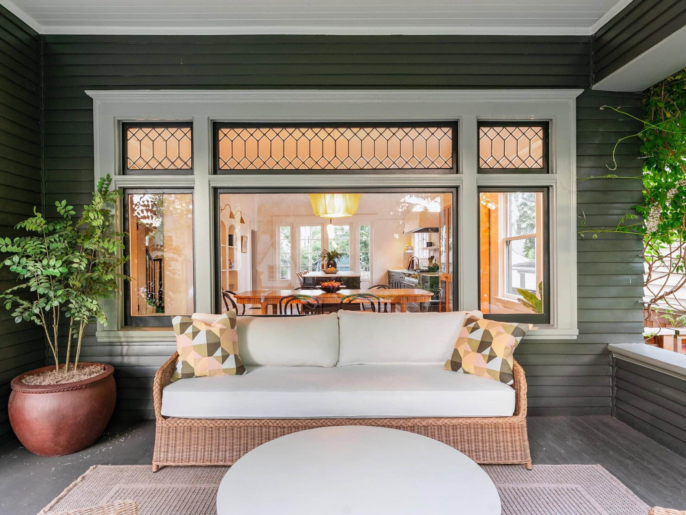 A cozy porch with a wicker sofa, patterned pillows, a round white table, and a large potted plant. The porch overlooks a well-lit dining room through a window with decorative glass panes.