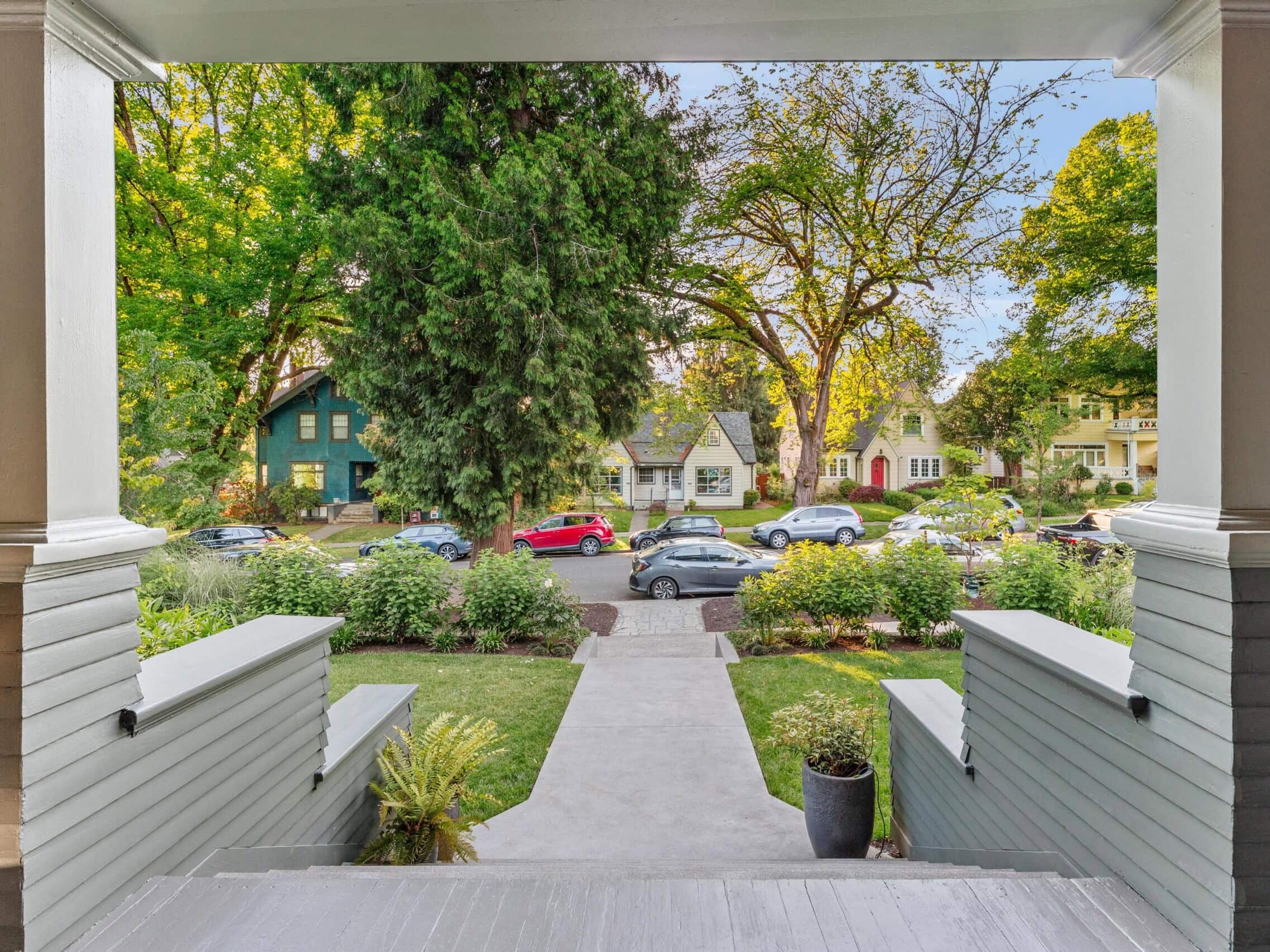 View from a front porch looking out at a tree-lined neighborhood street with houses, parked cars, and lush greenery. Steps and pathway lead from the porch to the sidewalk.