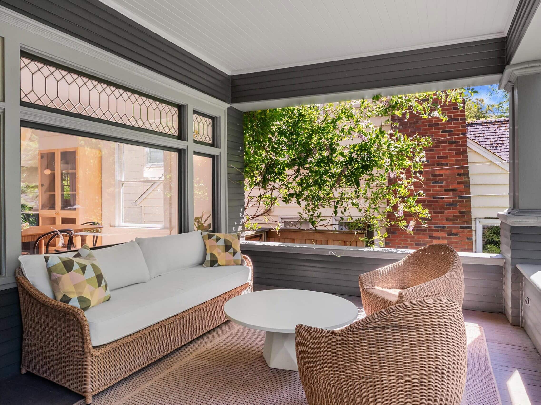 A cozy covered porch with a wicker sofa, two wicker chairs, a round white coffee table, and a rug. The porch overlooks a brick house and greenery, with sunlight streaming in through open sides.