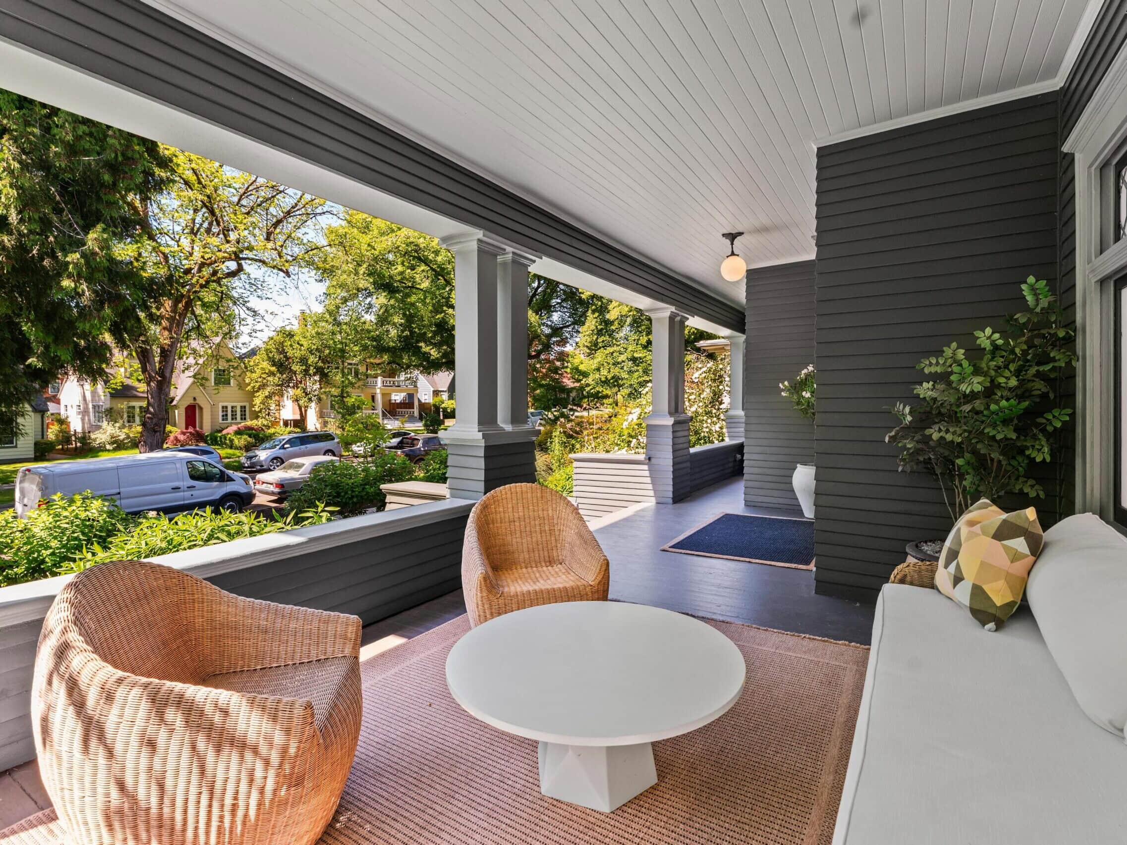 A covered porch with wicker chairs, a white round table, and a white sofa. Trees and parked cars line the street in front of houses, visible beyond the porch railing on a sunny day.
