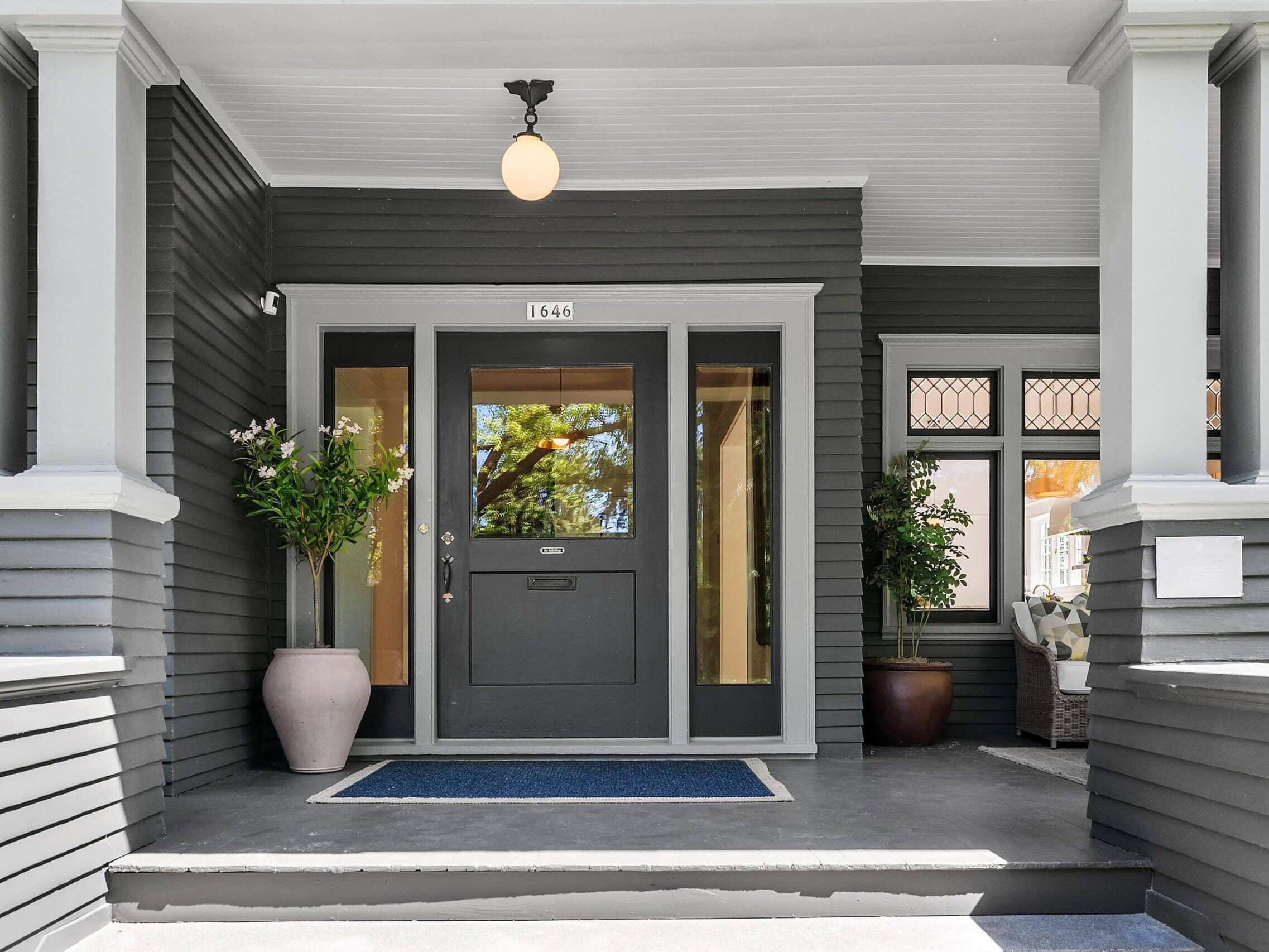 A gray house entrance with a central door and sidelights, a porch with columns, two potted plants, a blue doormat, and a round porch light above the door displaying the number 1646.