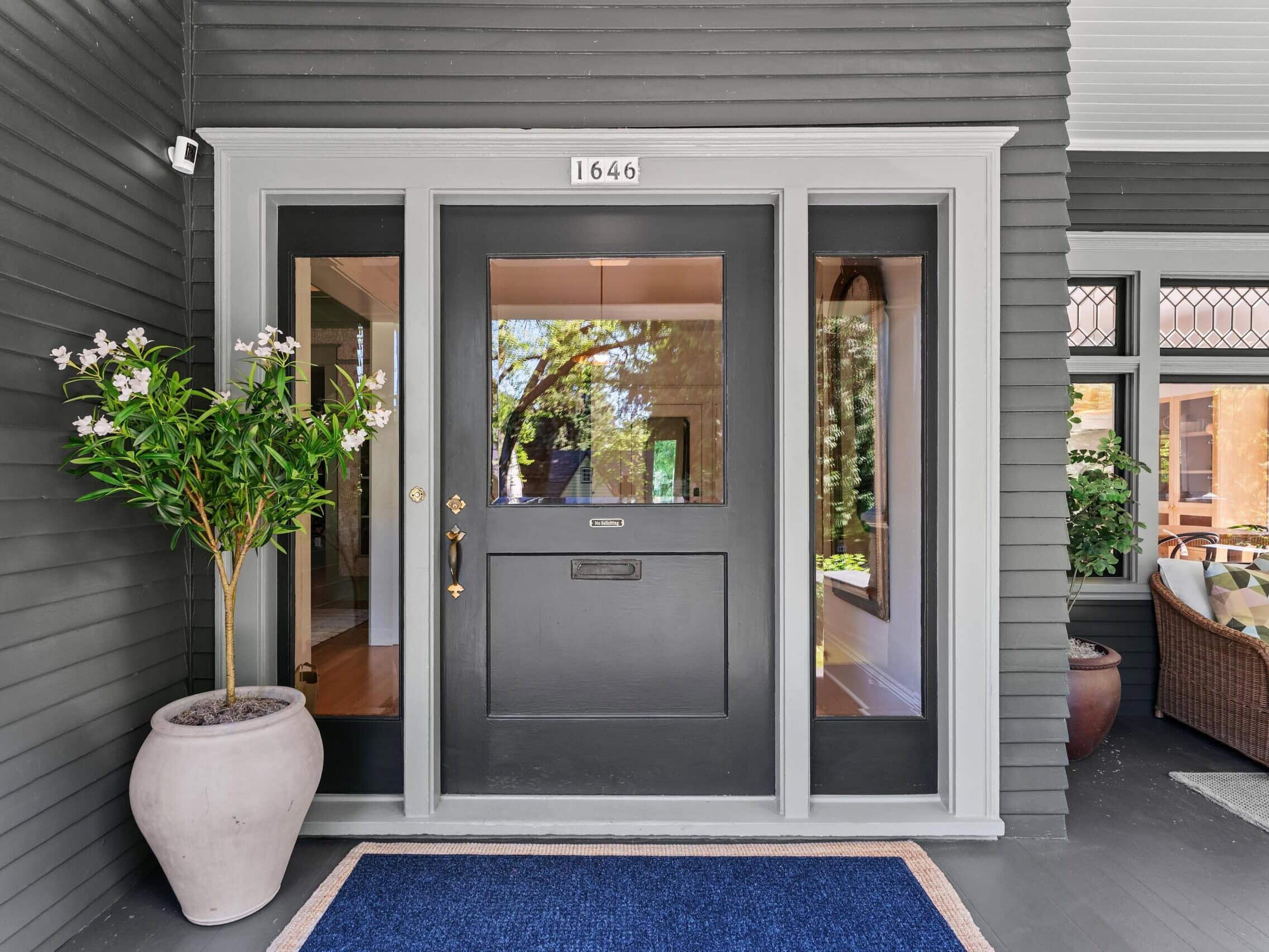 A modern black front door with glass panels, flanked by large windows, a potted plant on the left, and a blue welcome mat on a gray porch. The house number 1646 is displayed above the door.
