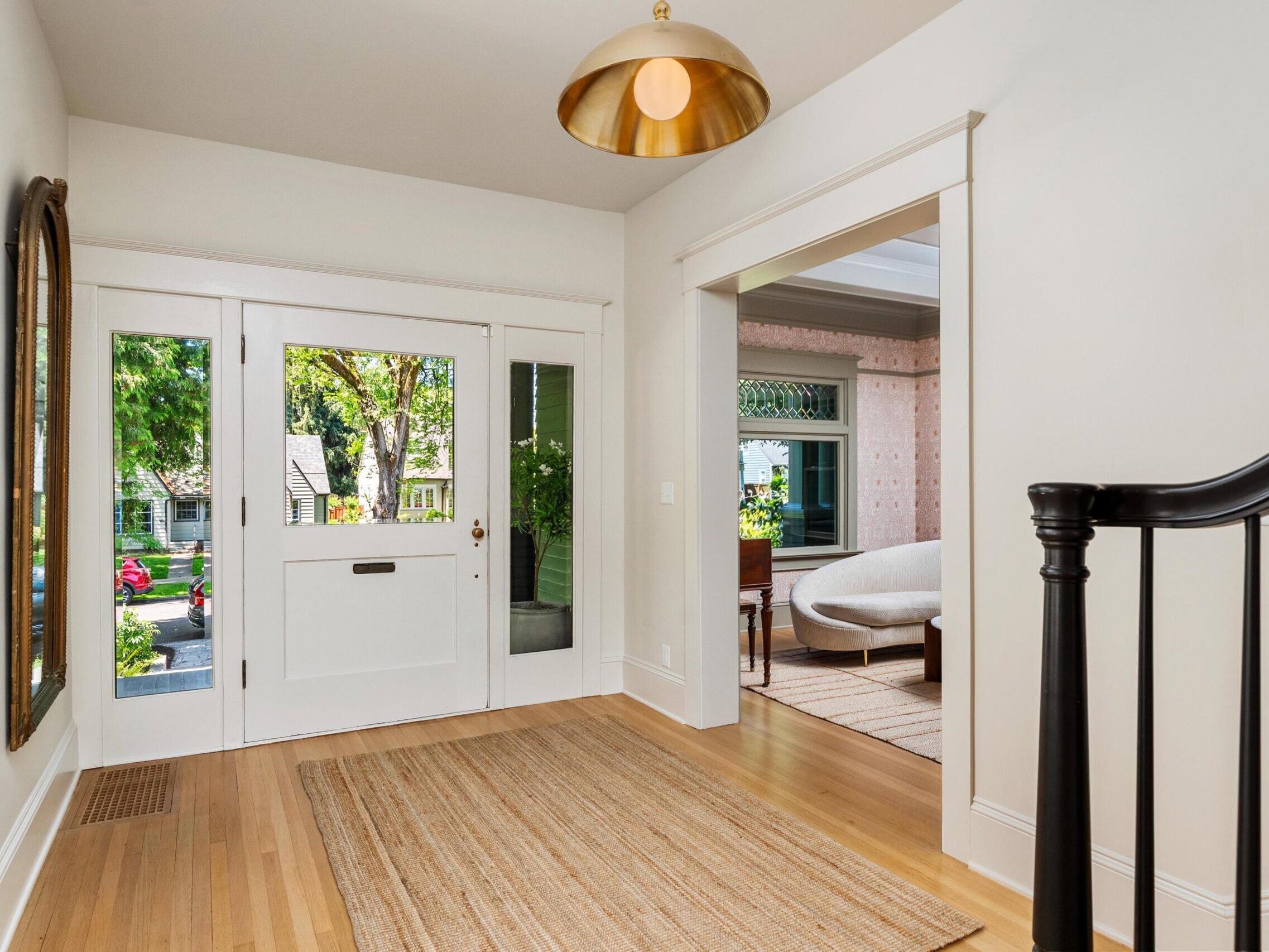 A bright entryway with light wood floors, a woven rug, white walls, and a large windowed front door. A round mirror and brass light fixture decorate the space, with a view into a cozy, furnished room beyond.