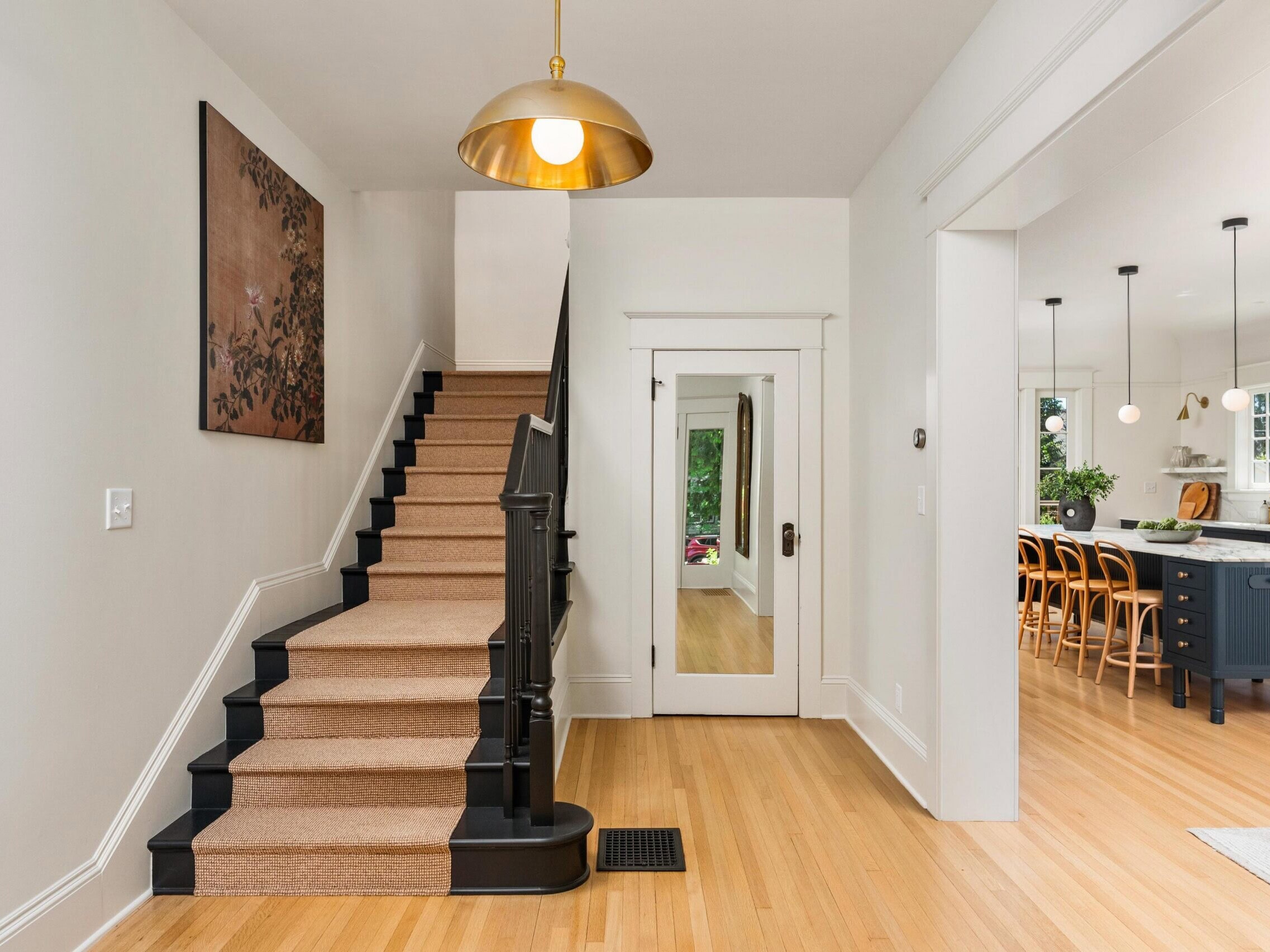 Bright entryway with hardwood floors, a carpeted staircase with black railings, a white door with a mirror, a modern gold pendant light, and a view into a kitchen with barstools and pendant lights.