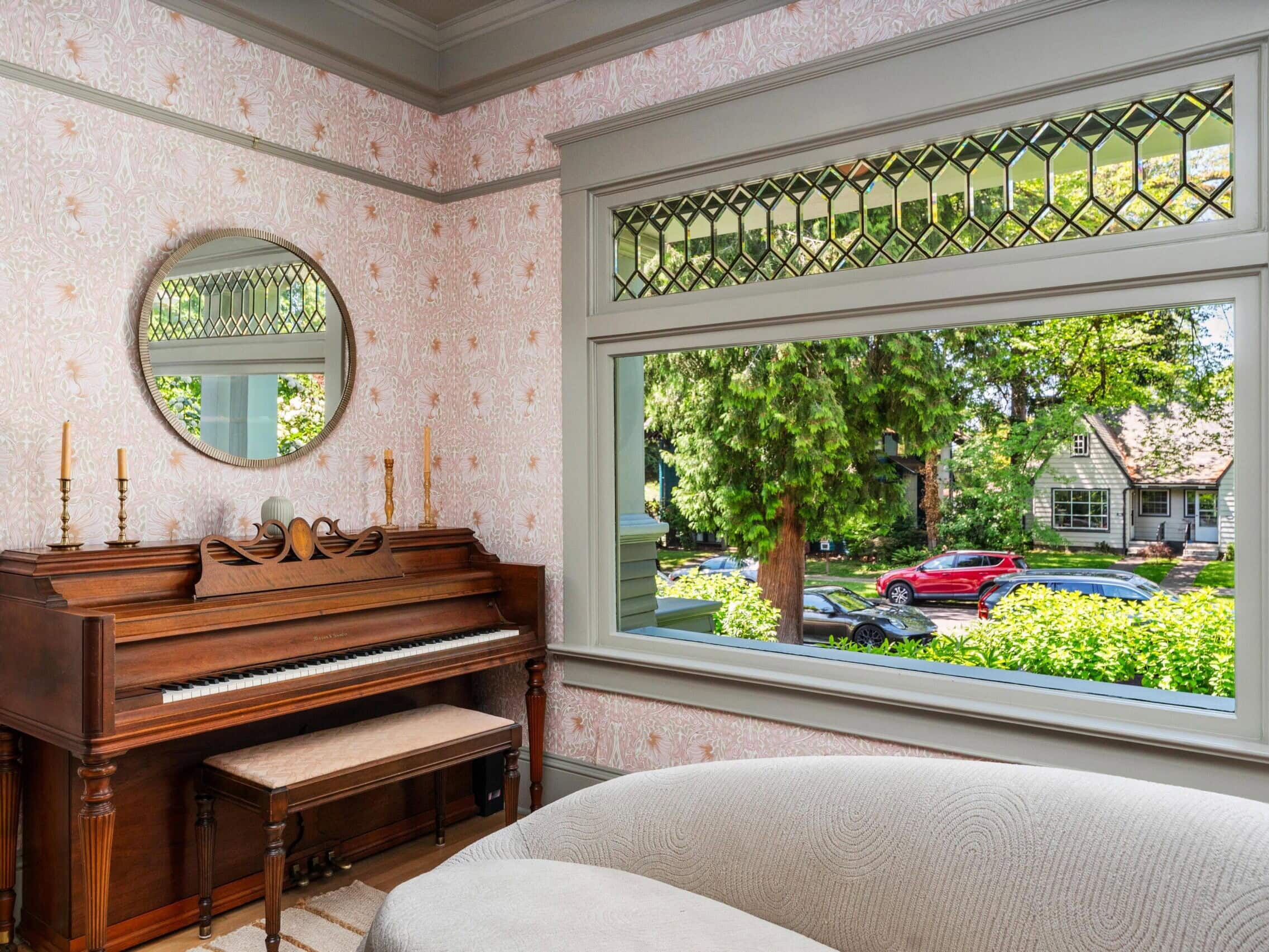 A cozy room with a wooden piano and bench, two candlesticks, a round mirror above the piano, floral wallpaper, a large window with stained glass trim, and a view of trees and houses outside.