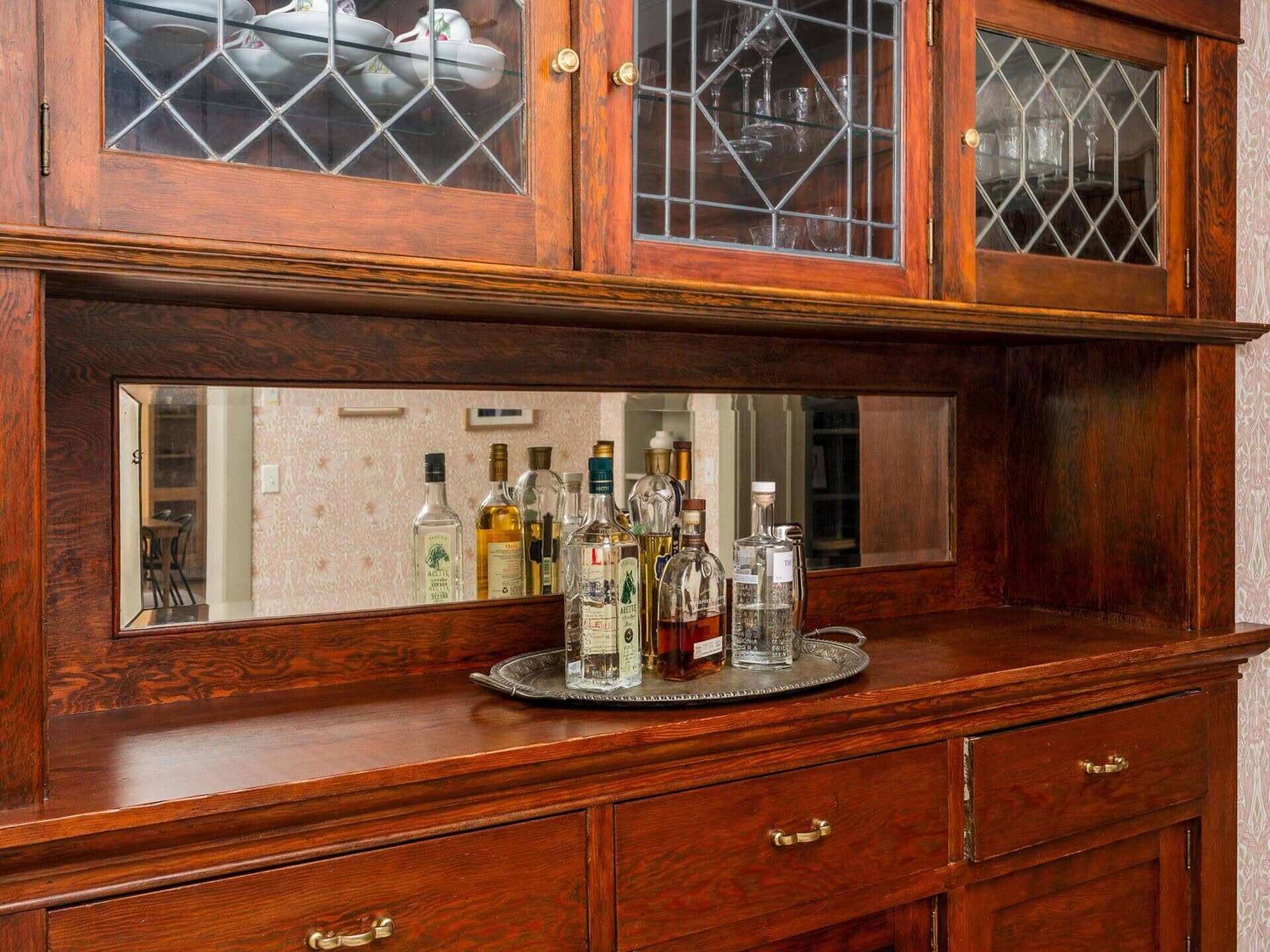 A wooden sideboard with glass cabinets on top, displaying dishes. The countertop holds a tray with various liquor bottles and glasses, with a mirror behind them. The furniture has brass handles and sits against patterned wallpaper.