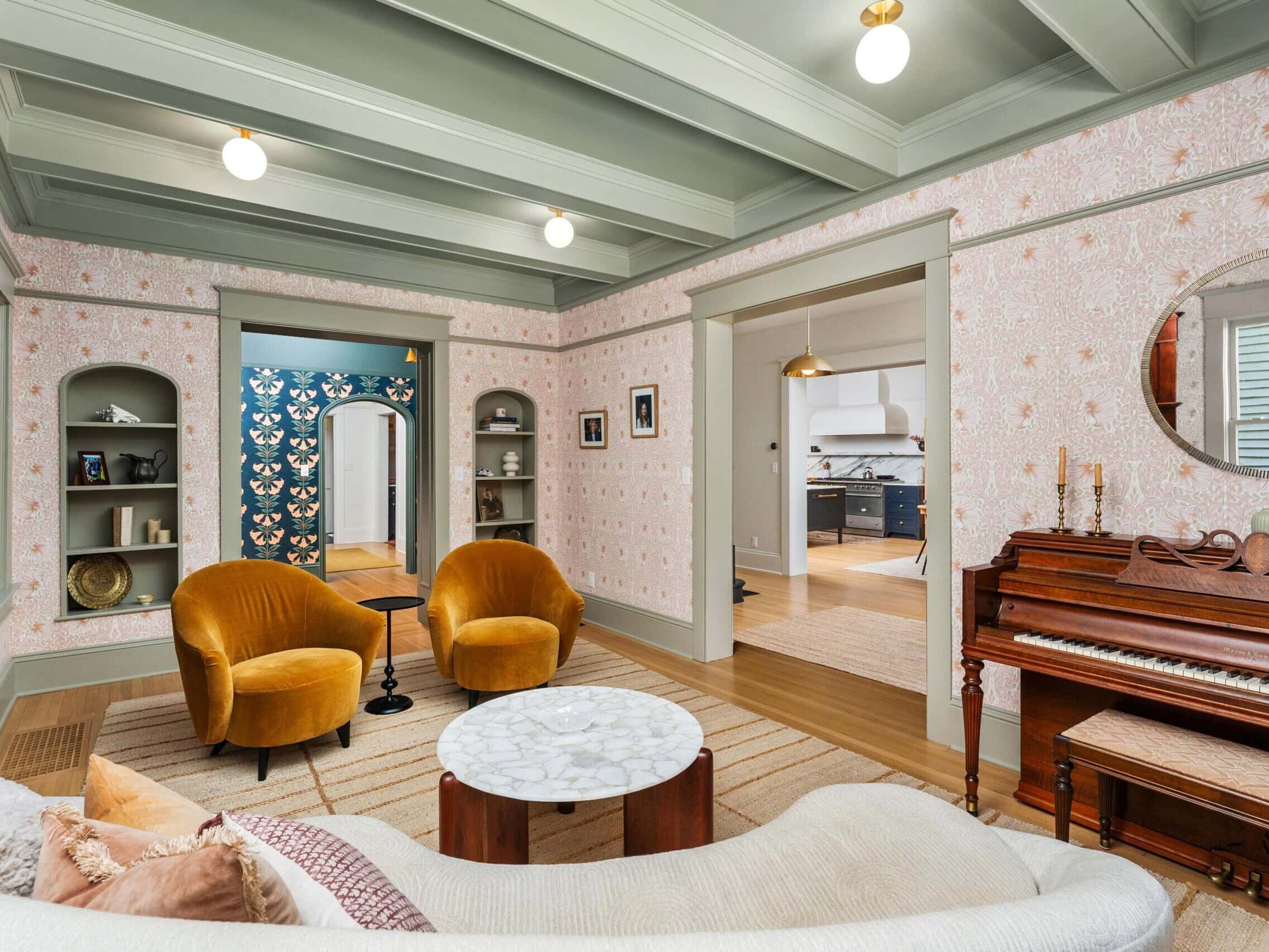 A cozy living room with a vintage piano, mustard yellow armchairs, a curved sofa, a round marble coffee table, built-in shelves, floral wallpaper, and views into a kitchen and a hallway with blue floral wallpaper.