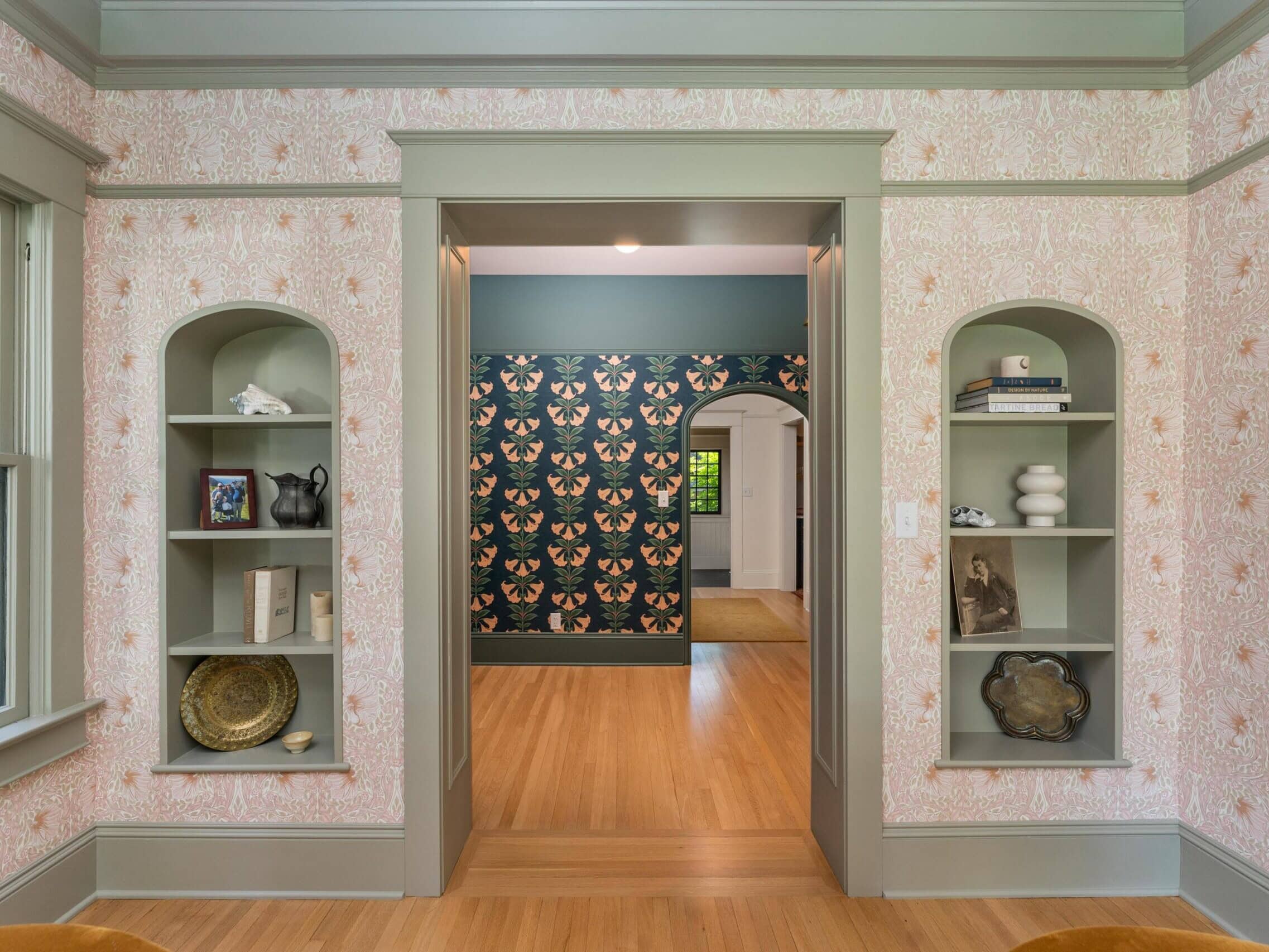A room with built-in arched shelves on either side of a doorway, floral wallpaper, and light wood floors. Through the doorway, another room with butterfly-patterned wallpaper is visible.