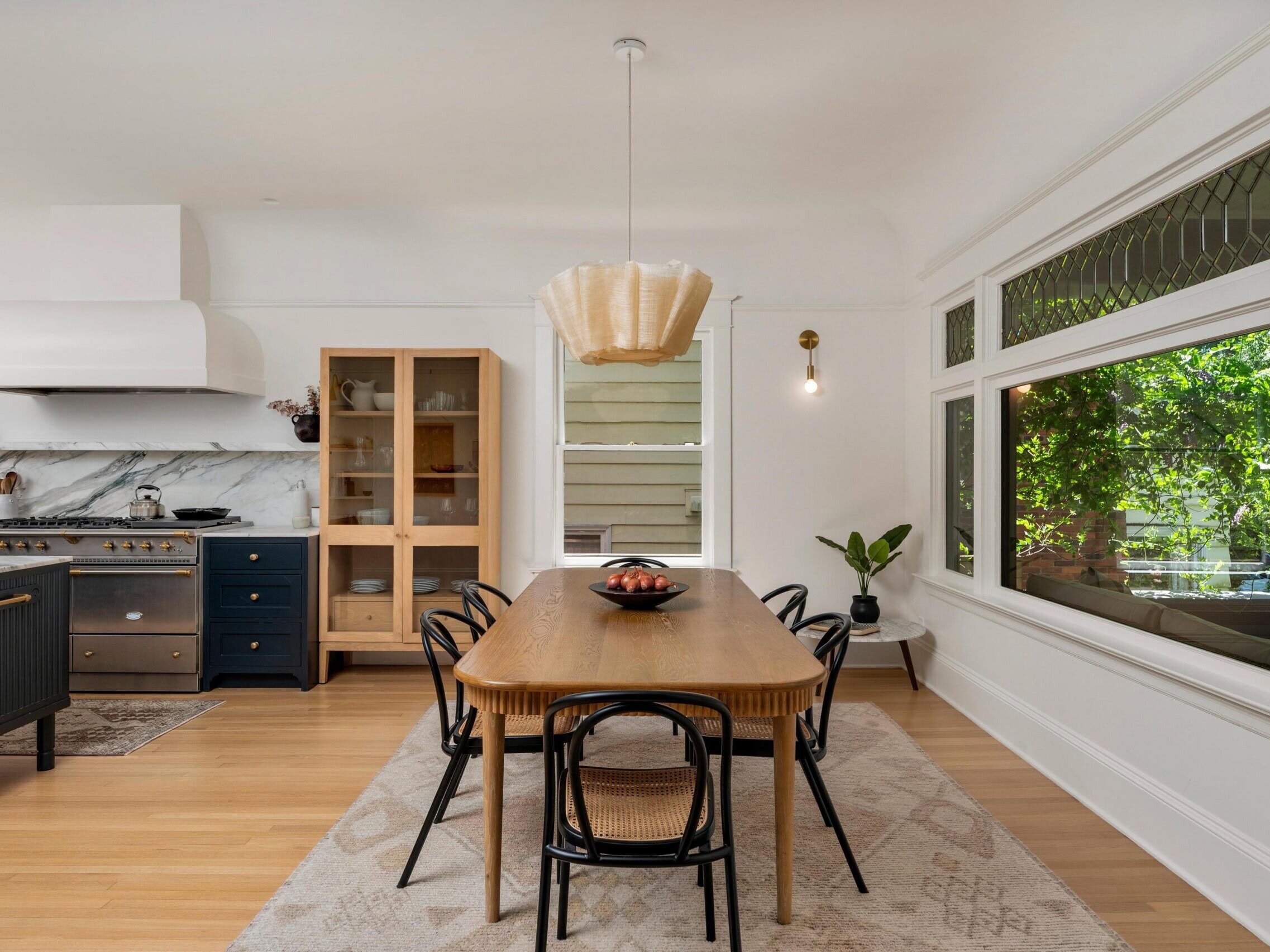 Bright, modern kitchen and dining area with a wooden table, four black chairs, pendant light, black stove, glass-front cabinet, large window, and indoor plant. Natural light fills the room.