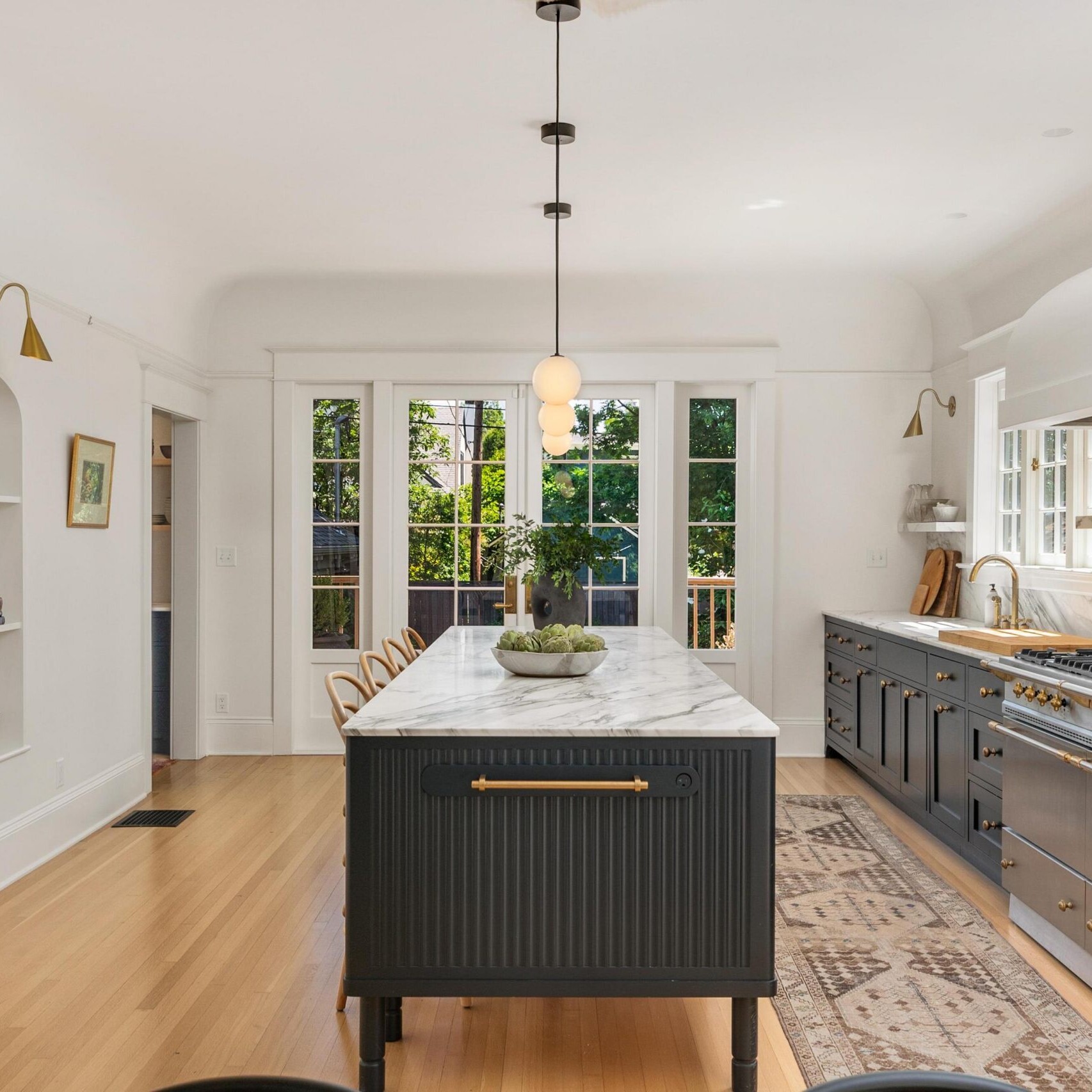Elegant kitchen with a large marble island, dark cabinets, gold hardware, and open shelving. Glass doors let in natural light. Modern light fixtures and greenery complete the bright, stylish space—just one example from a top real estate agent in Portland.