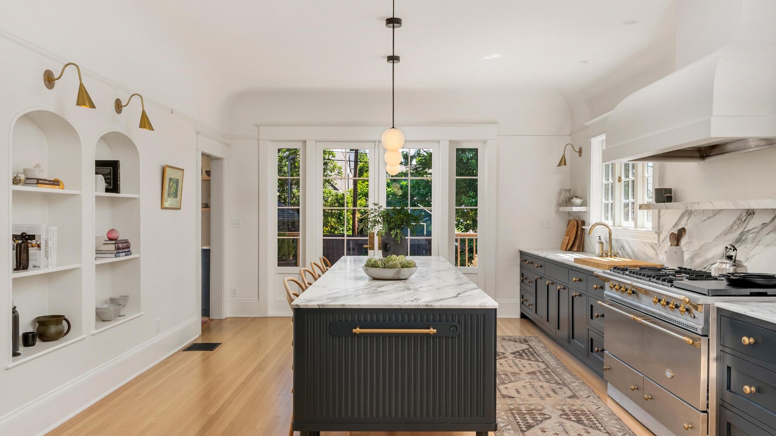 Elegant kitchen with a large marble island, dark cabinets, gold hardware, and open shelving. Glass doors let in natural light. Modern light fixtures and greenery complete the bright, stylish space—just one example from a top real estate agent in Portland.