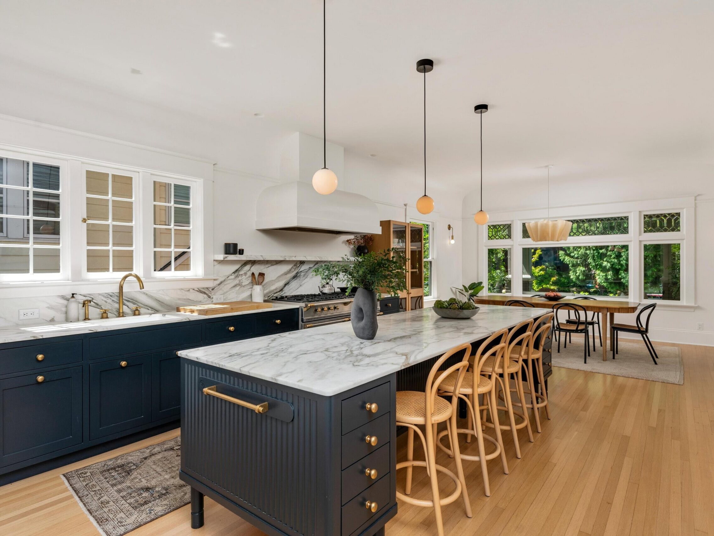 Modern kitchen with a large marble island, wooden bar stools, dark blue cabinets, pendant lighting, and an adjacent dining area with large windows overlooking greenery.