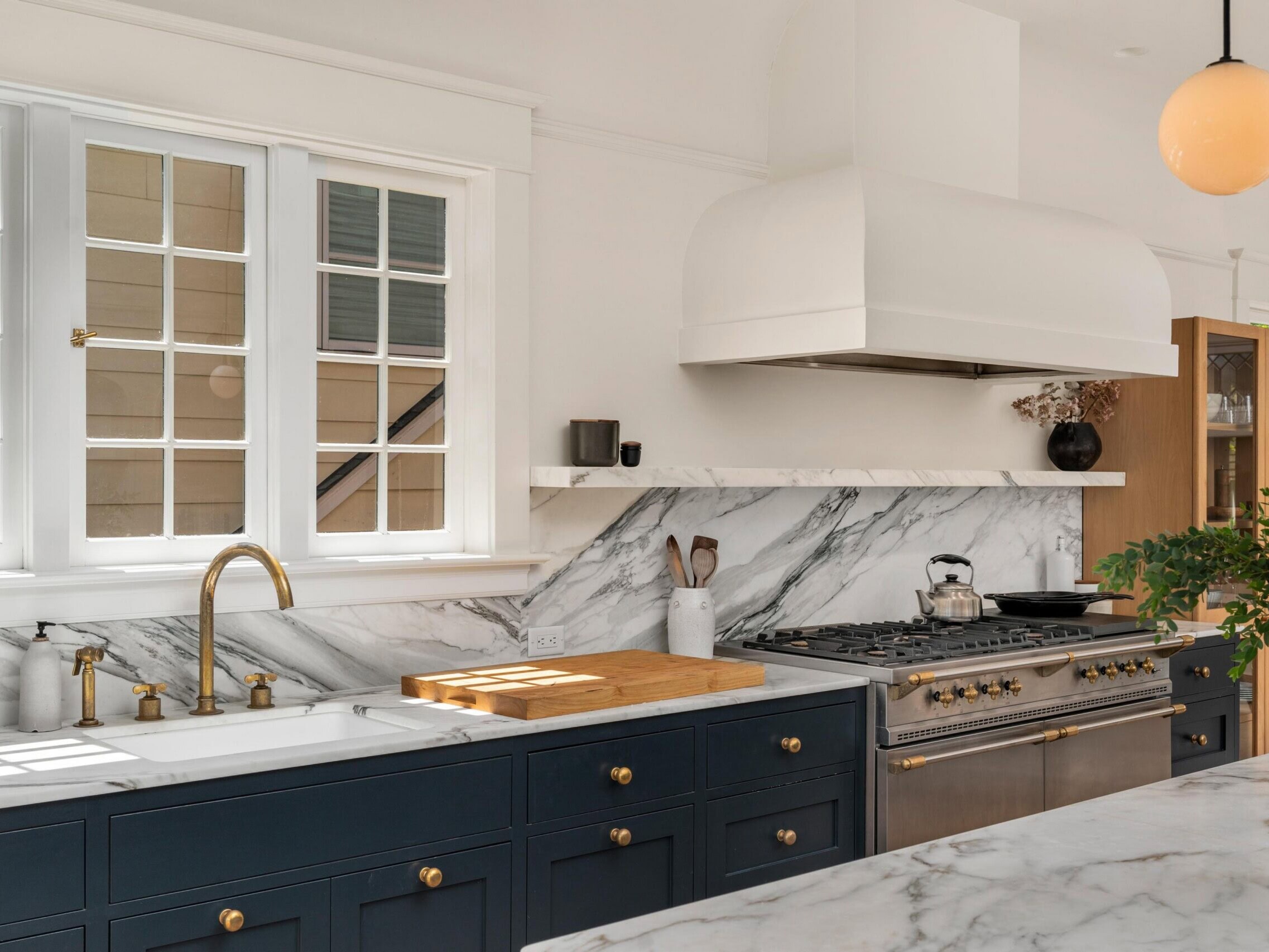 Modern kitchen with navy cabinets, brass hardware, a white farmhouse sink, marble countertops and backsplash, stainless steel gas range, white hood, open shelf with decor, and large window letting in natural light.