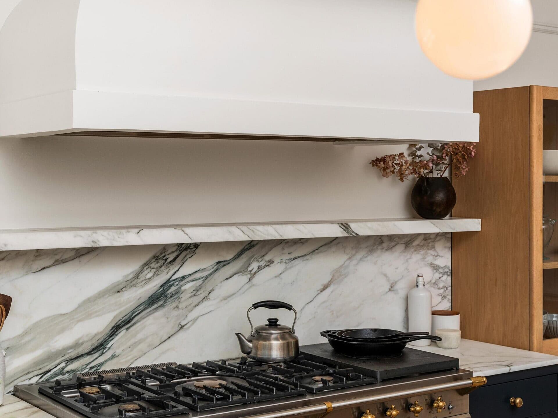 A modern kitchen features a marble backsplash, a stainless steel gas stove with brass accents, a kettle, a skillet, and minimalist white and wood cabinetry. A round pendant light hangs from the ceiling.