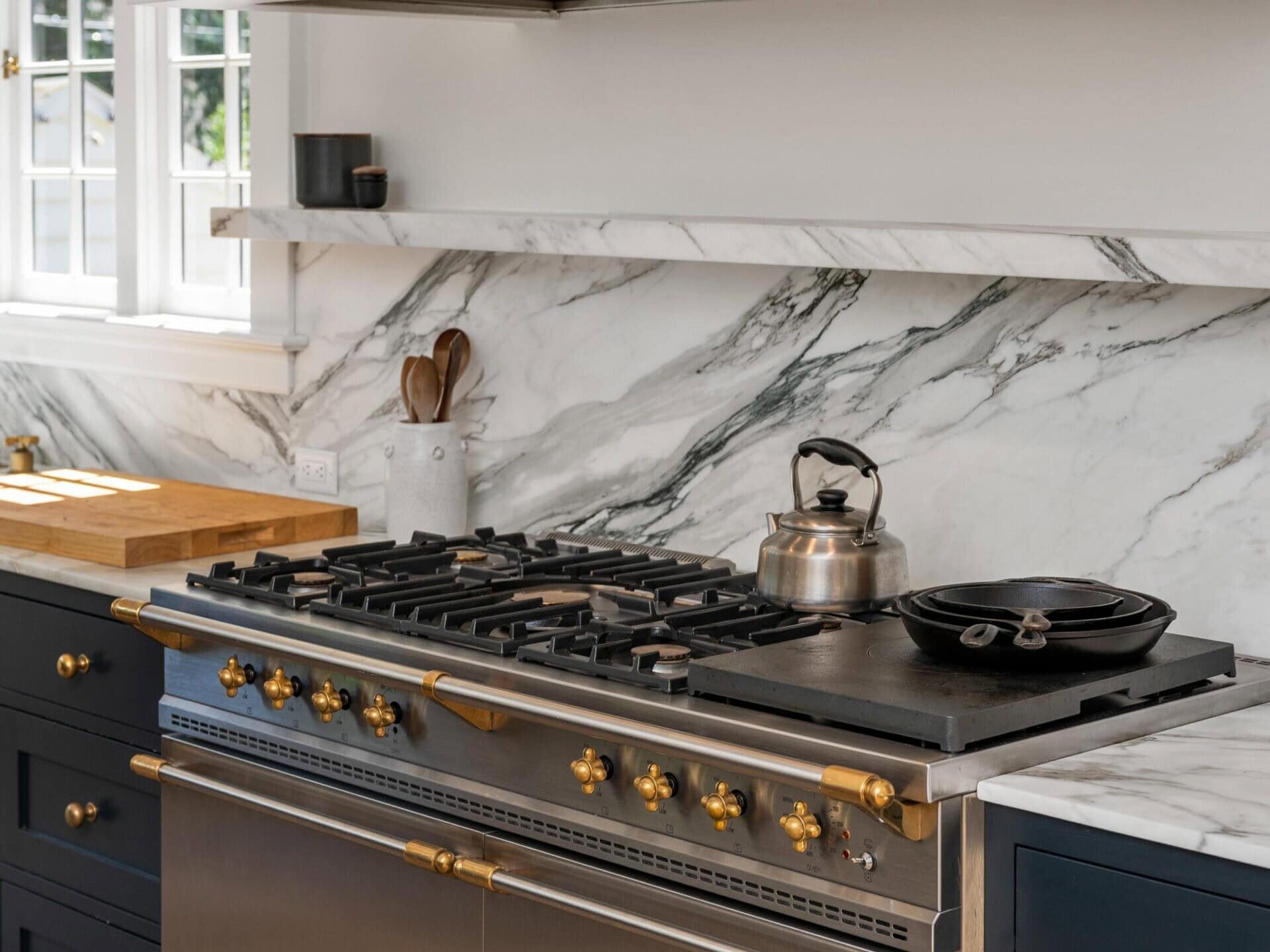 A modern kitchen features a large stainless steel gas stove with brass accents, a silver kettle, and a pot on the burners. The backsplash is white marble with gray veining, and there are dark blue cabinets below.