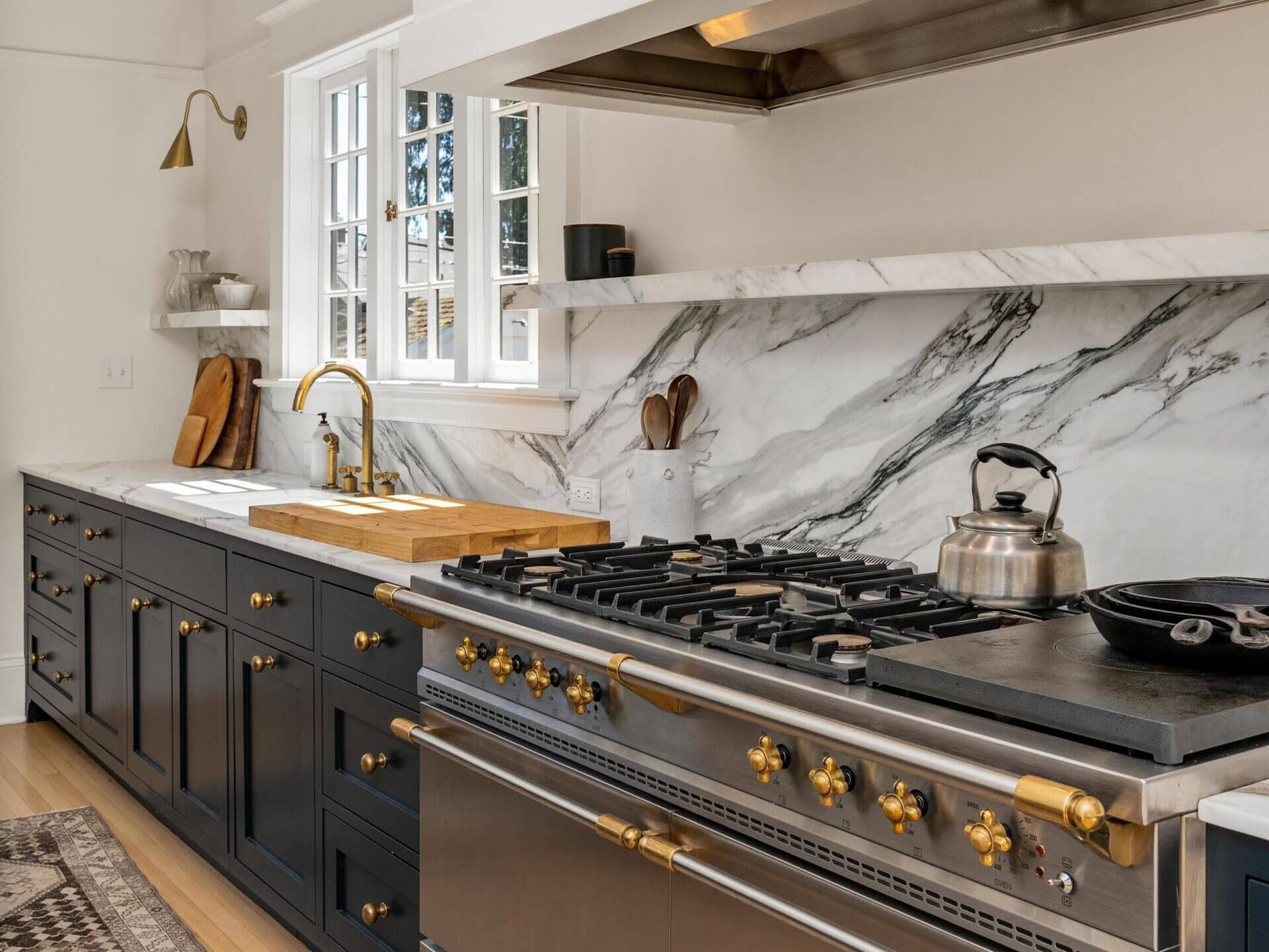A modern kitchen featuring dark blue cabinets with brass handles, a marble backsplash, a large stainless steel stove with brass knobs, a farmhouse sink, gold faucet, open shelving, and a patterned rug on a light wood floor.
